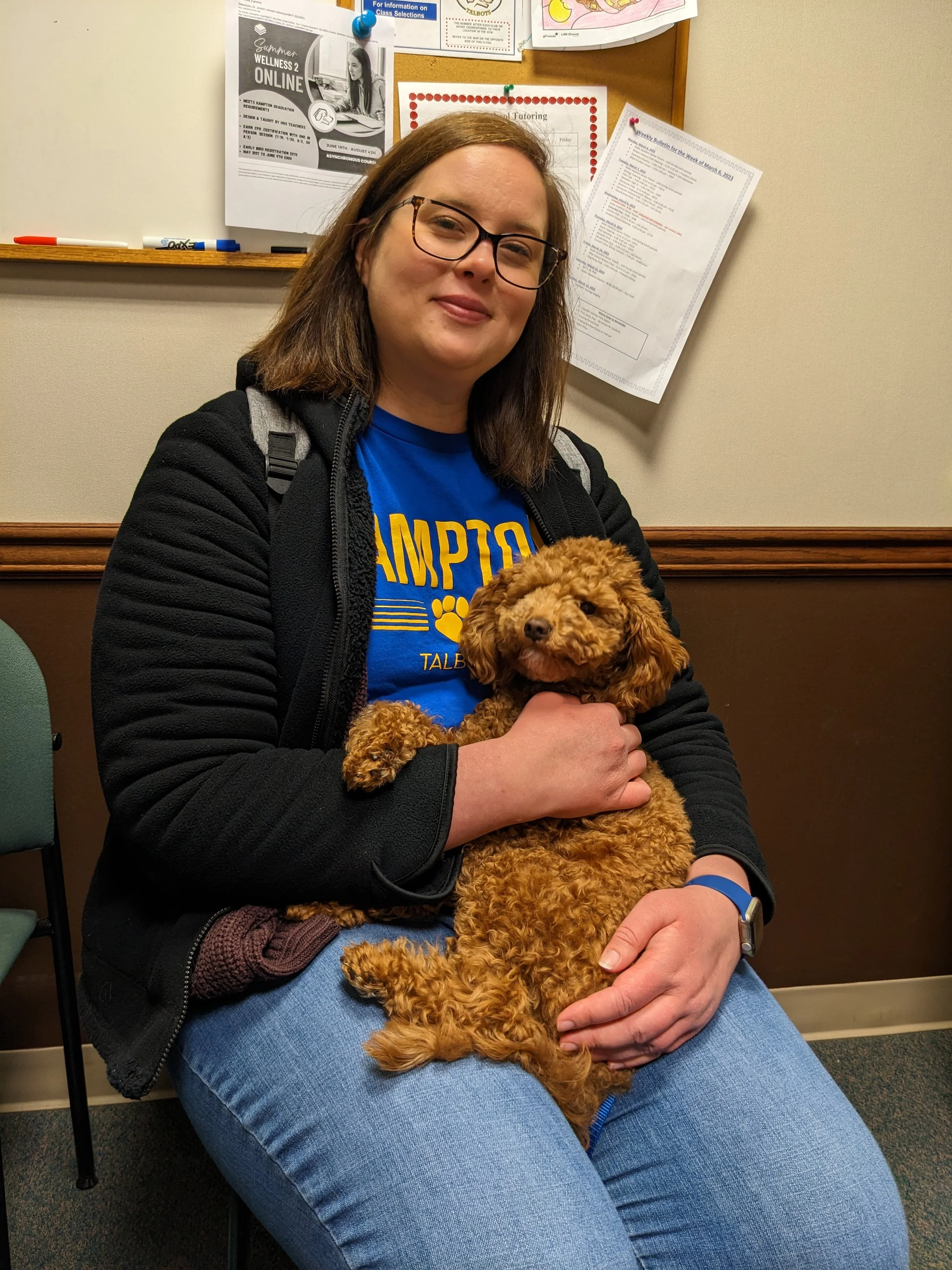 A woman with glasses and brown hair sitting on a chair, holding a small curly-haired brown dog on her lap. The woman is wearing a blue shirt and a black jacket, and is smiling.