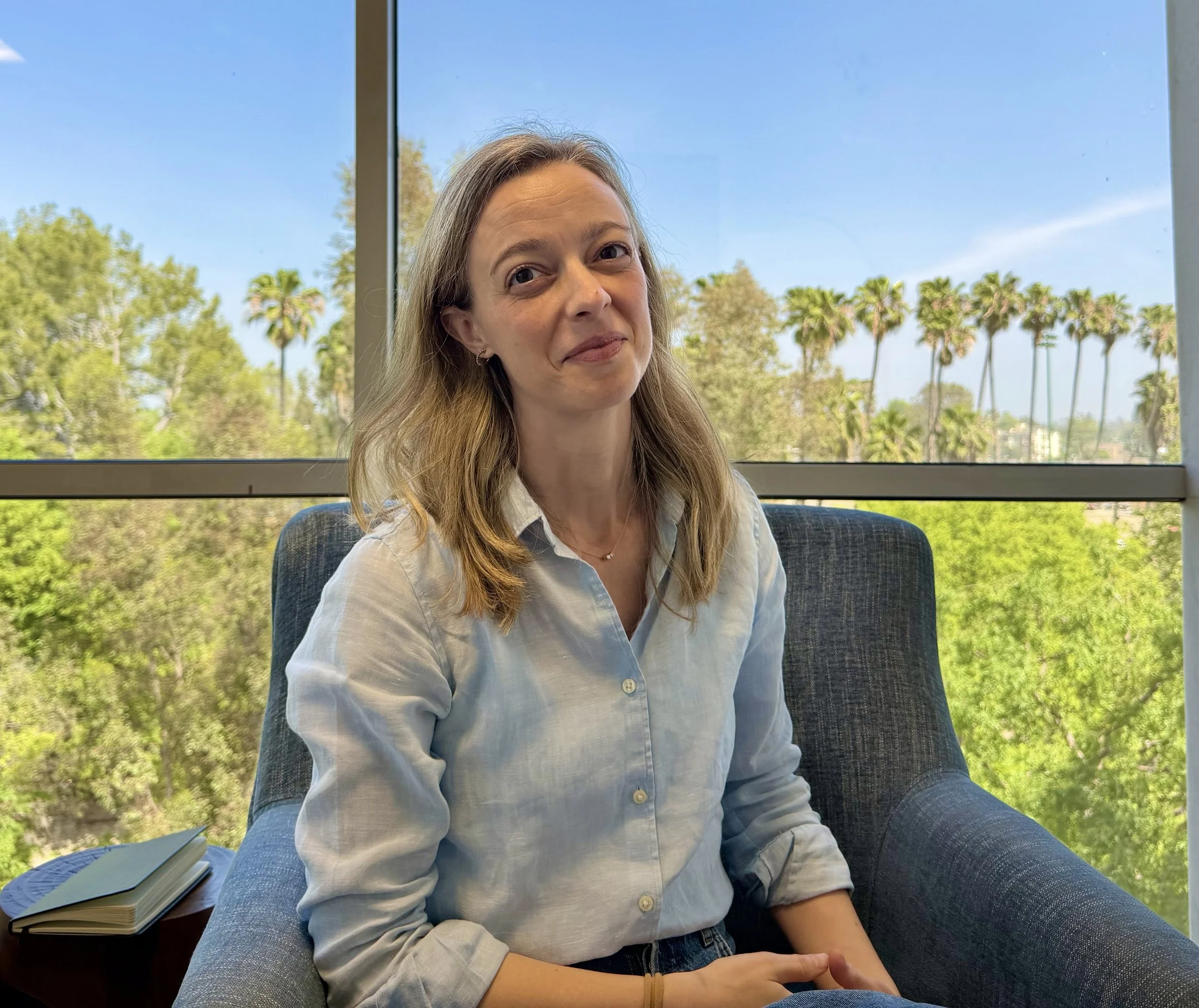 A woman who is a therapist with a compassionate smile, with light brown hair, wearing a white button-up shirt, sitting in a gray armchair near a large window with a view of trees and palm trees outside.