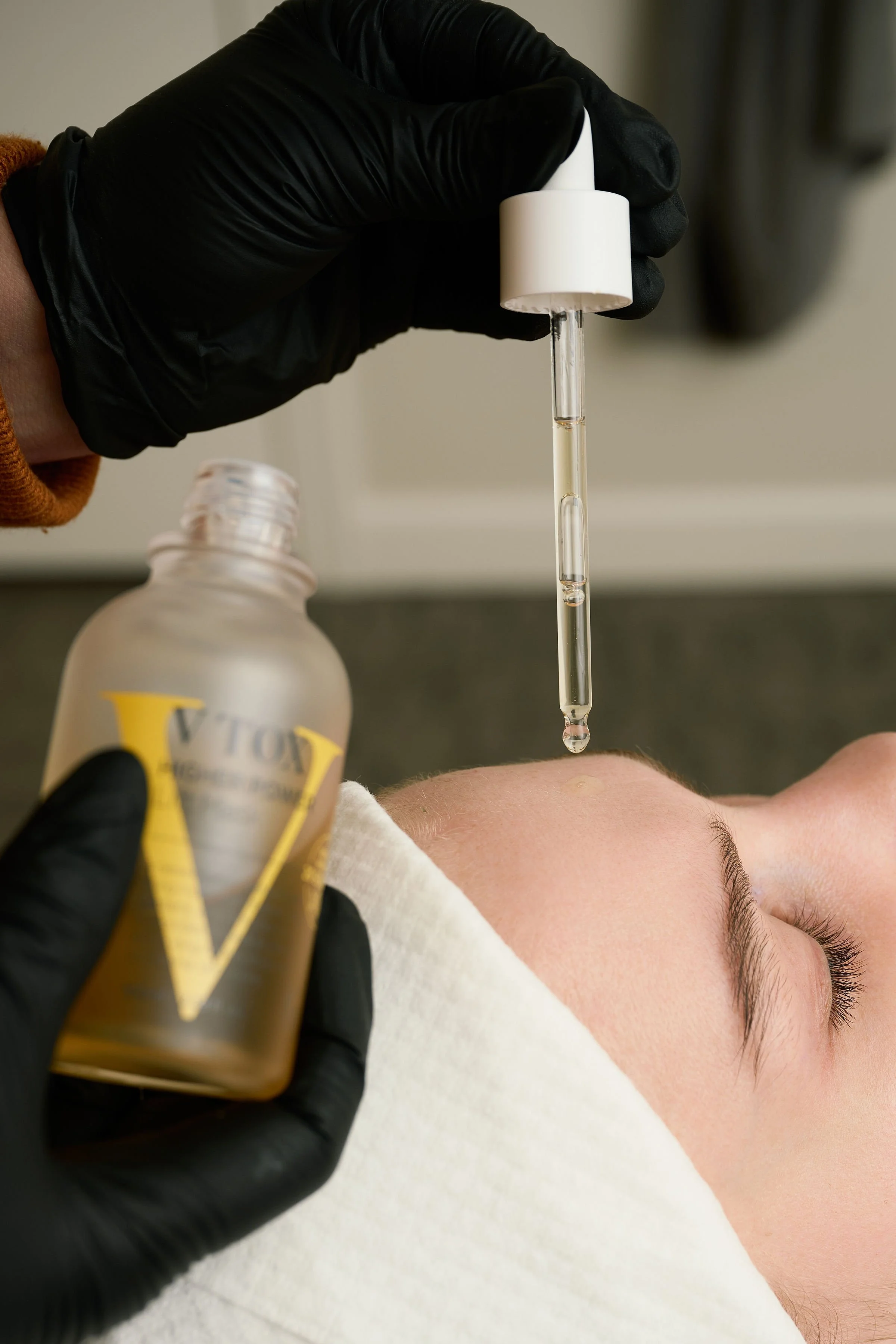 A person receiving a facial treatment using a dropper with a skincare serum or oil, held by a practitioner wearing black gloves, with a bottle of serum in hand and a patient lying down with a white facial mask or towel.