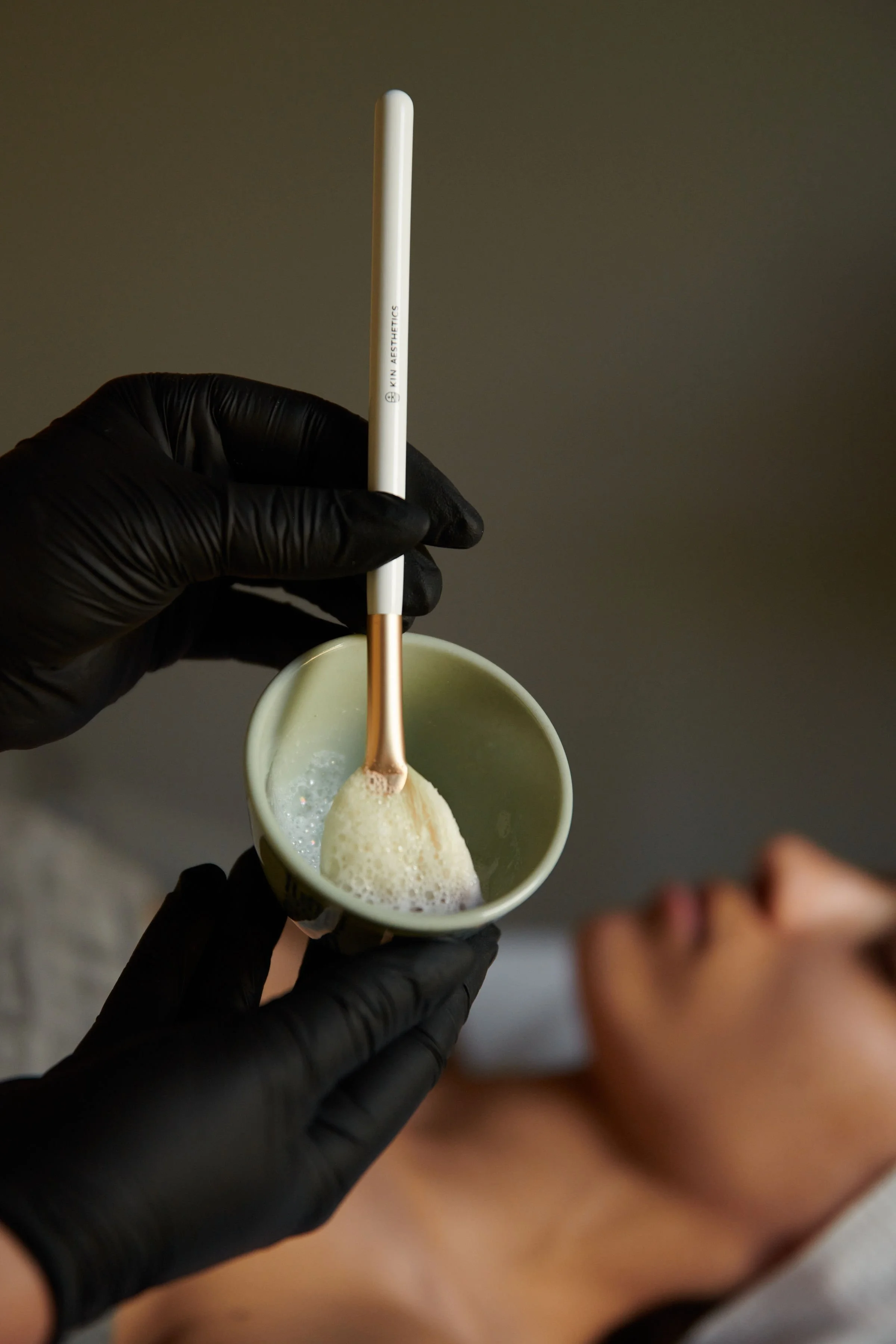 A person wearing black gloves holds a small cup with foam and a tool inside it, while a woman receives a facial treatment in the background.