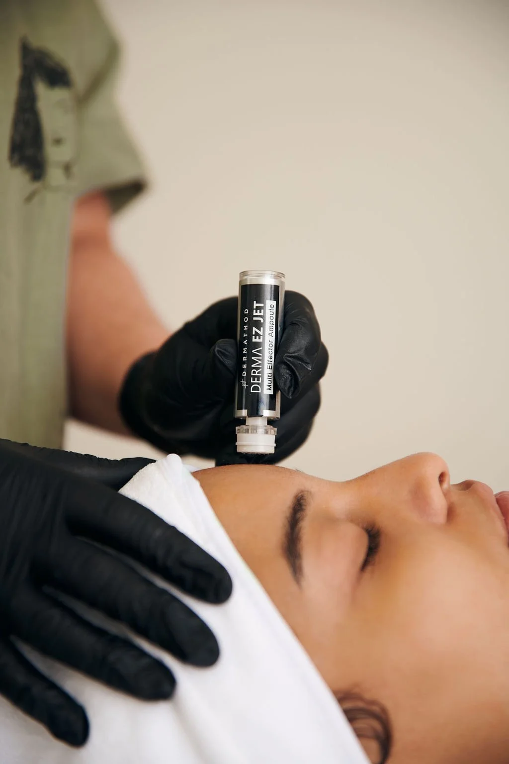 A person receiving a cosmetic treatment using a Dermal EZ Jet device, with a technician wearing black gloves applying the treatment to the person's cheek.