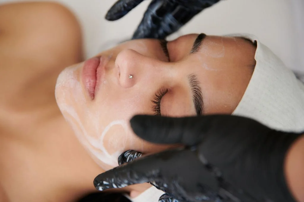A woman lying down with her eyes closed, receiving a skincare treatment from an esthetician wearing black gloves, with a headband and facial masks applied.