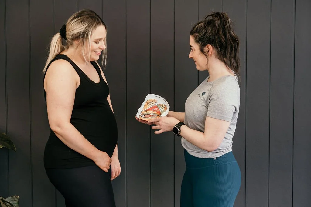 A pregnant woman smiling as she looks at a diagram of the pelvic floor held by a woman in workout attire.