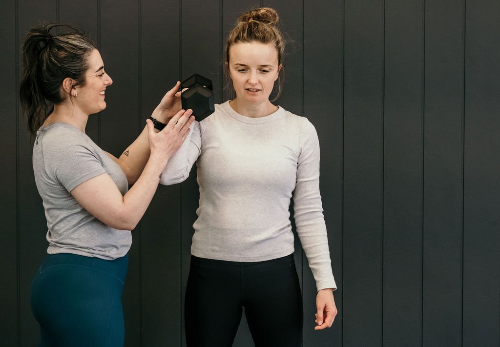 Two women working out together, with one holding a small black dumbbell while the other expects to lift it.