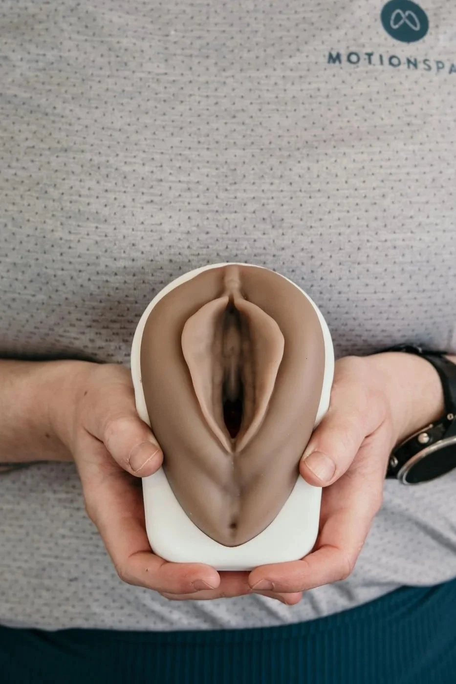 A person holding a realistic female genitalia model. The person is wearing a gray shirt with a logo and a black watch.