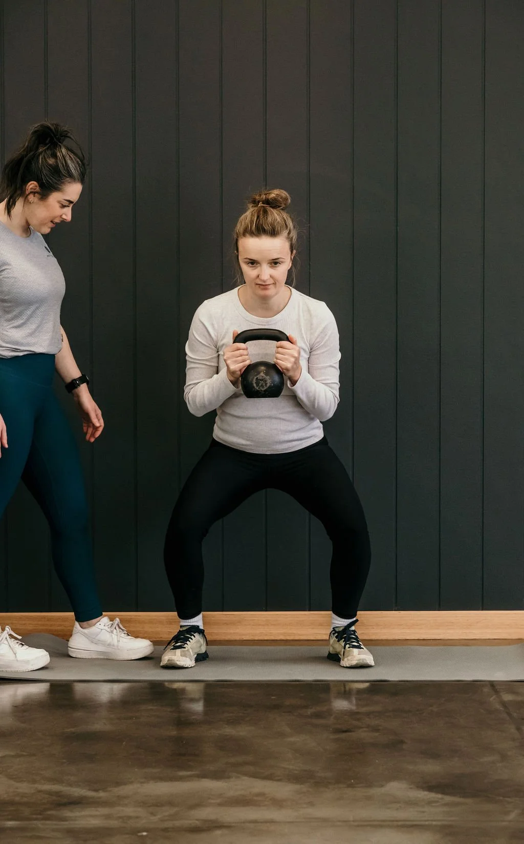 A woman holding a kettlebell in a squat stance in a gym, with a trainer observing beside her.