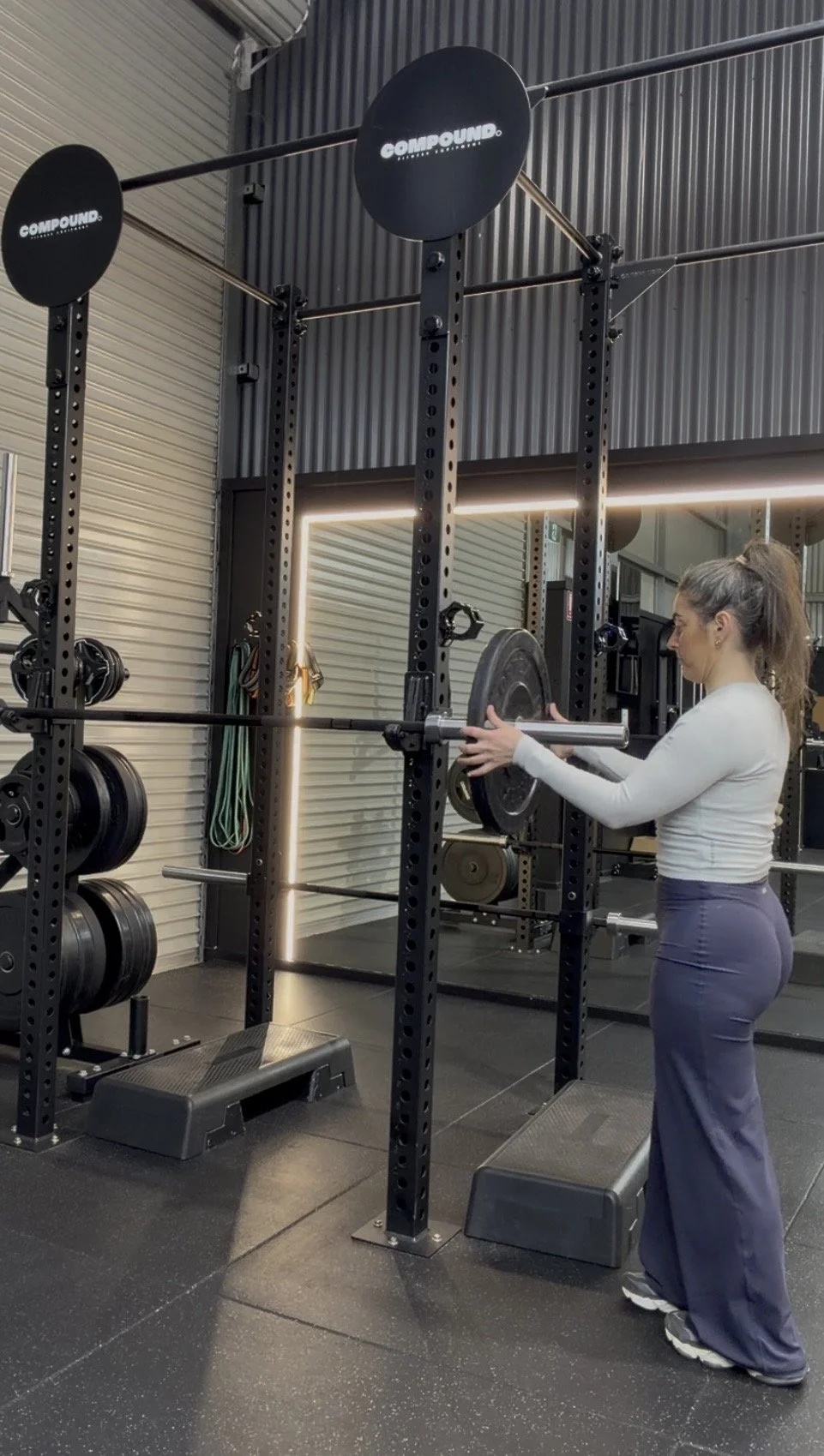 A woman lifting a barbell in a gym with black weights and a squat rack.