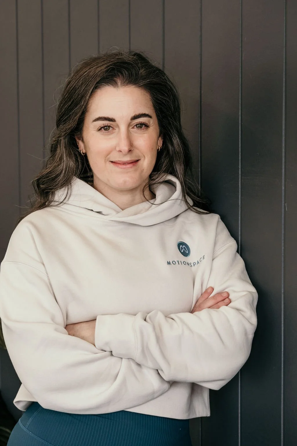 A young woman with dark hair and light skin, wearing a white hoodie with a logo and the word 'MOTIONSPACE,' standing with her arms crossed against a dark wooden-paneled wall, smiling slightly at the camera.