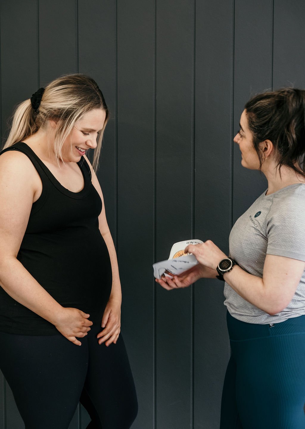 A woman in a gray workout shirt shows a model of the pelvic floor, while another woman in black workout attire laughs and looks down, standing against a dark wall.