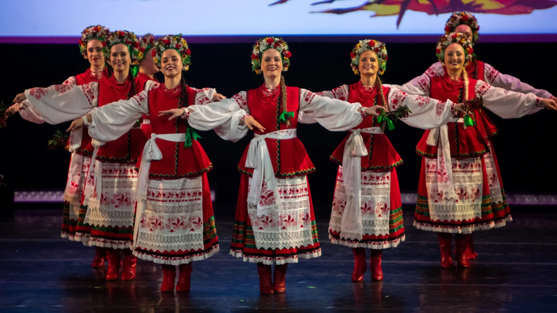 Female dancers from Rusalka Ukrainian Dance Ensemble performing in traditional red and white embroidered costumes with floral wreaths, celebrating Ukrainian culture on stage in Winnipeg.