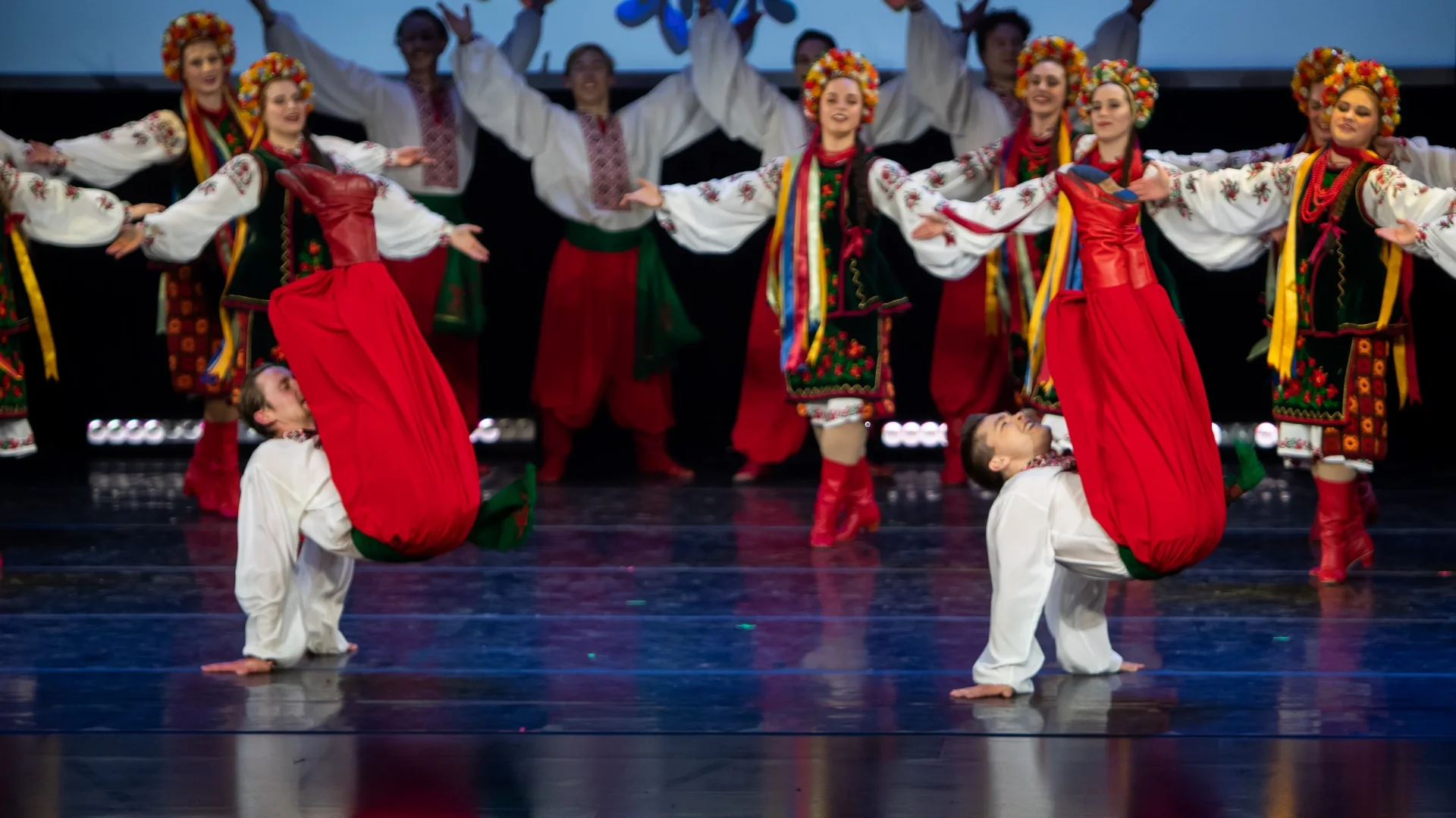 Male Rusalka Ukrainian dancers performing athletic acrobatic movements while female dancers in traditional embroidered costumes form a vibrant backdrop during a stage performance in Winnipeg.