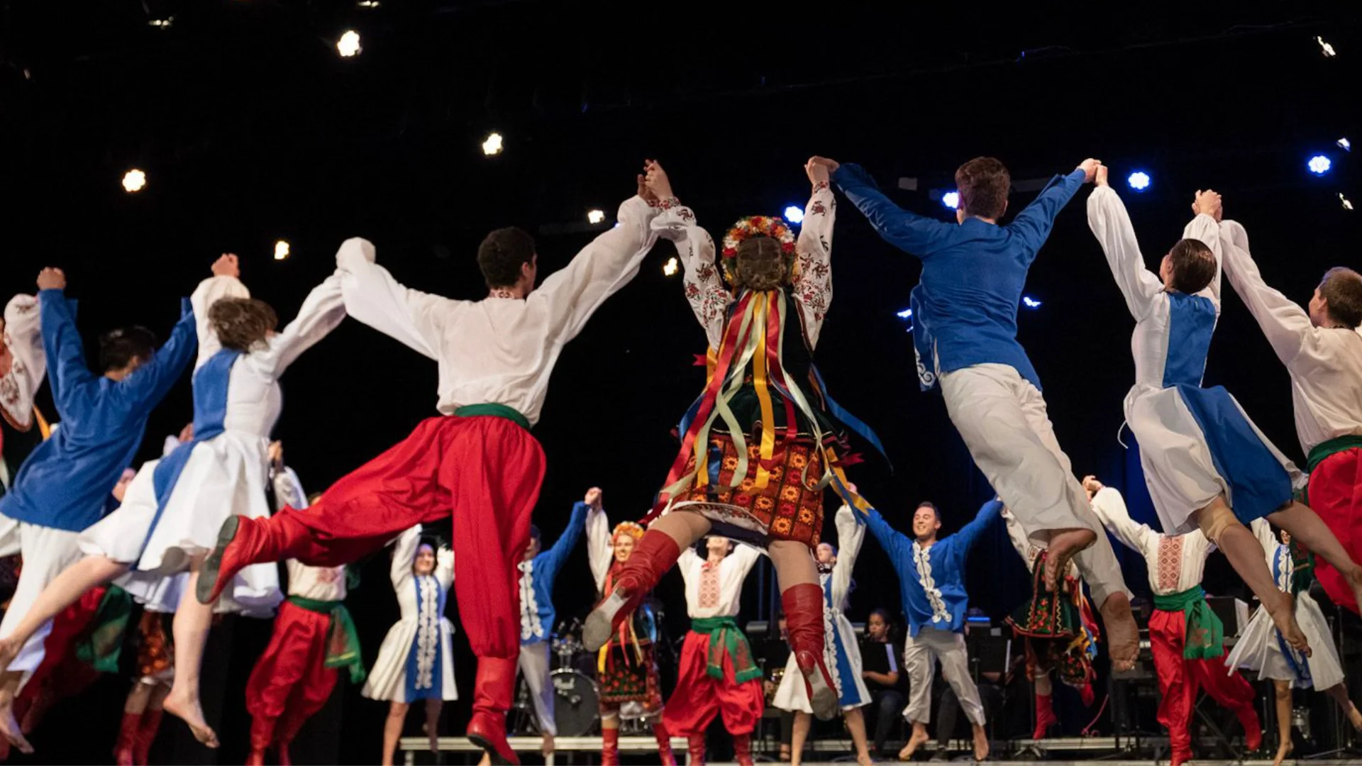 Rusalka Ukrainian Dance Ensemble captured mid-jump during a high-energy finale, with dancers in red, blue, and embroidered costumes holding hands on stage under theatrical lights in Winnipeg.