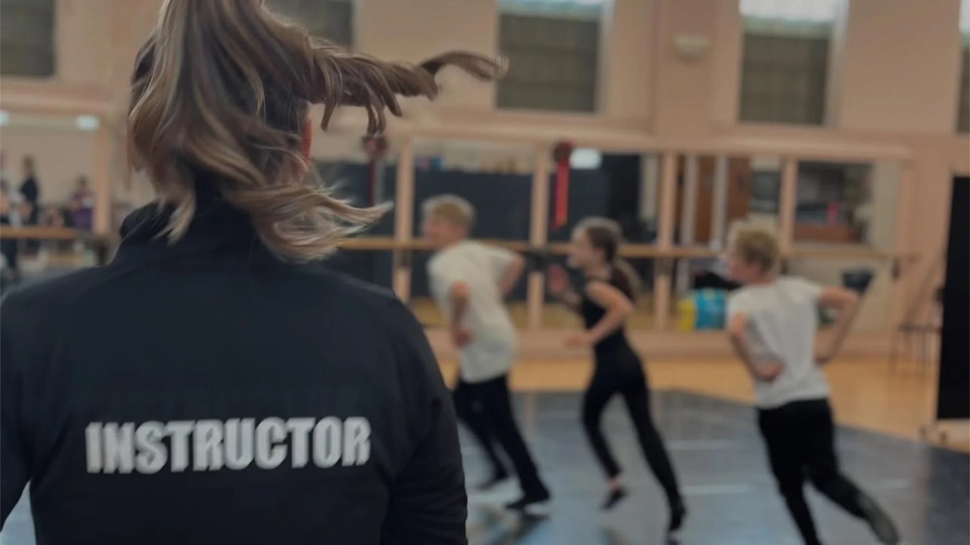 Dance instructor leading a class of children in a studio, with 'Instructor' visible on their shirt
