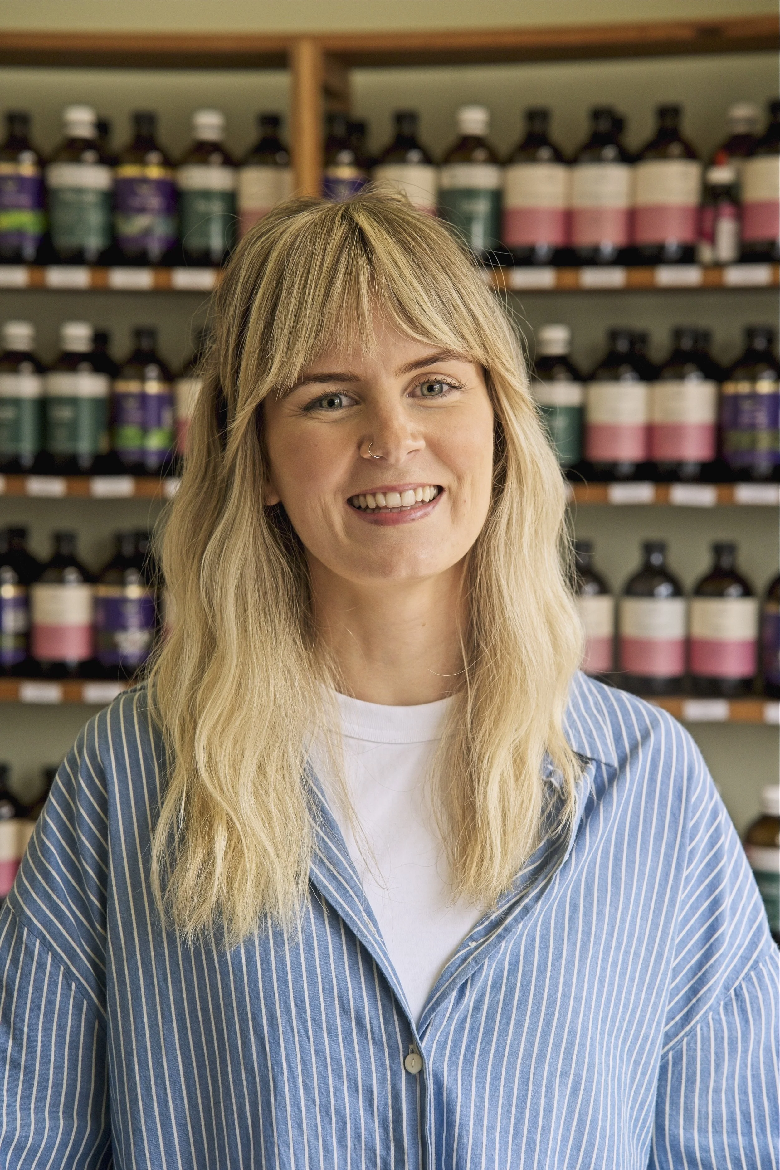 Headshot of Allie smiling warmly at the camera with shelves of herbal bottles in the background.