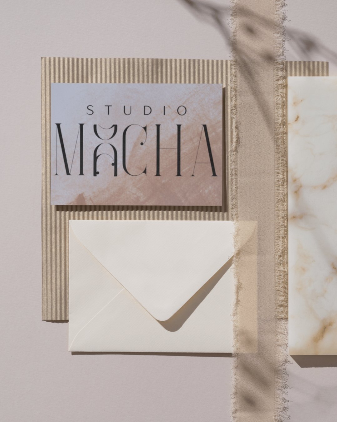 Flat lay of stationery items including a sign reading "Studio Màchia," cream-colored envelope, and textured paper with marble pattern on a light background.