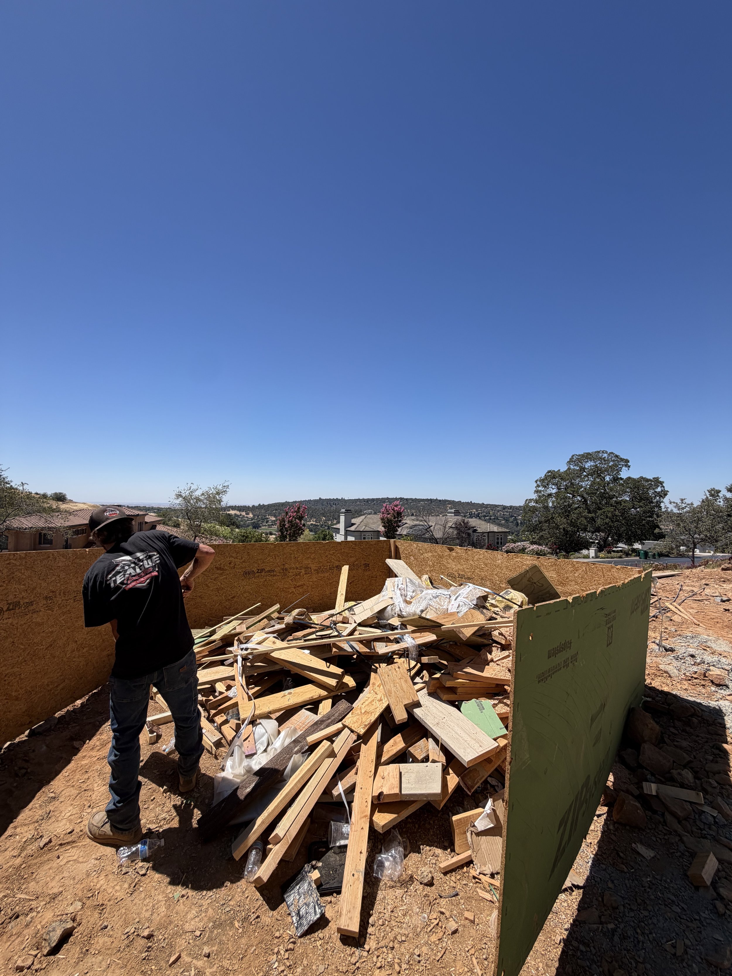 A construction worker wearing a Superman shirt, safety goggles, and a helmet working on a construction site with wood debris, cardboard boxes, and a green wall in a residential neighborhood under a clear blue sky.