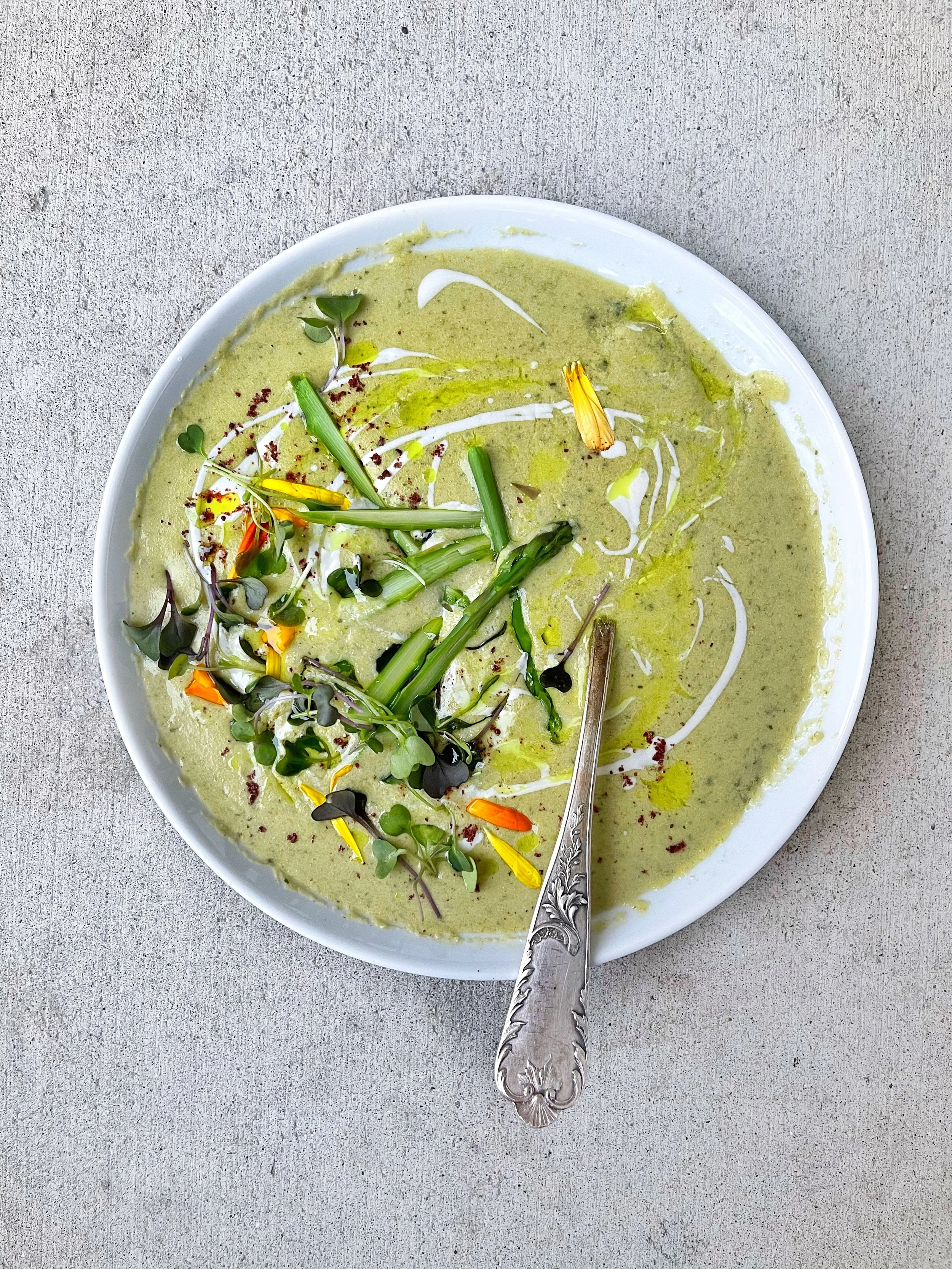 A white bowl filled with green soup garnished with microgreens, sliced green onions, yellow flower petals, and a metal spoon on a concrete surface.