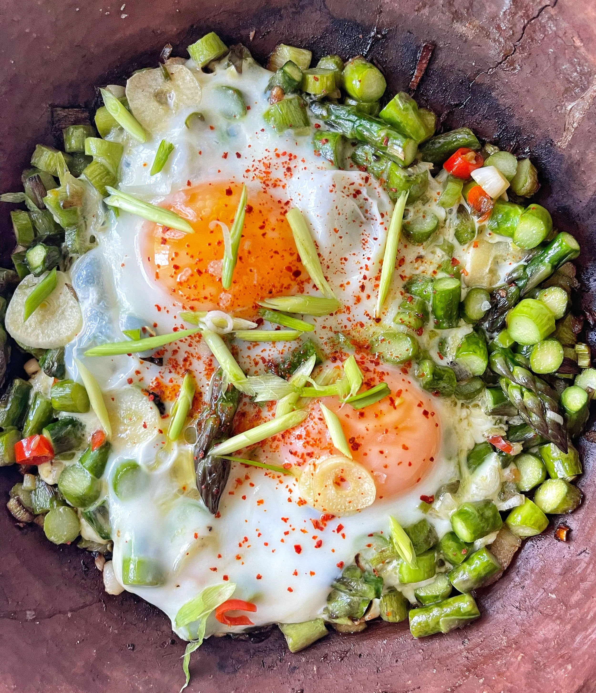 Fried eggs with asparagus, garlic, and chili flakes in a pan.