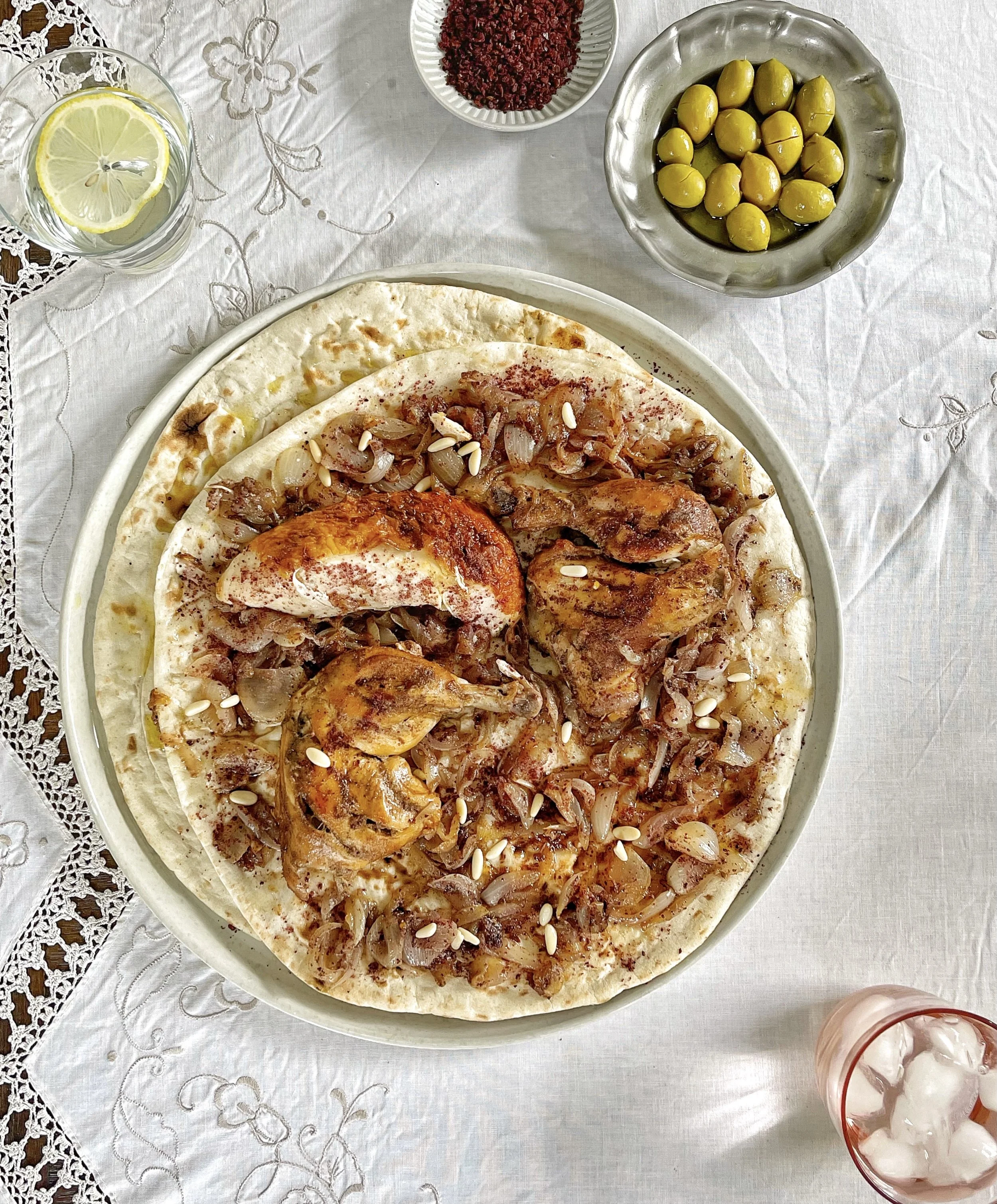 A plate of traditional Middle Eastern chicken musakhan, garnished with caramelized onions and pine nuts, served on flatbread. Accompanied by bowls of olives and ground spices, with a lemon water glass and an iced beverage on a decorative tablecloth.