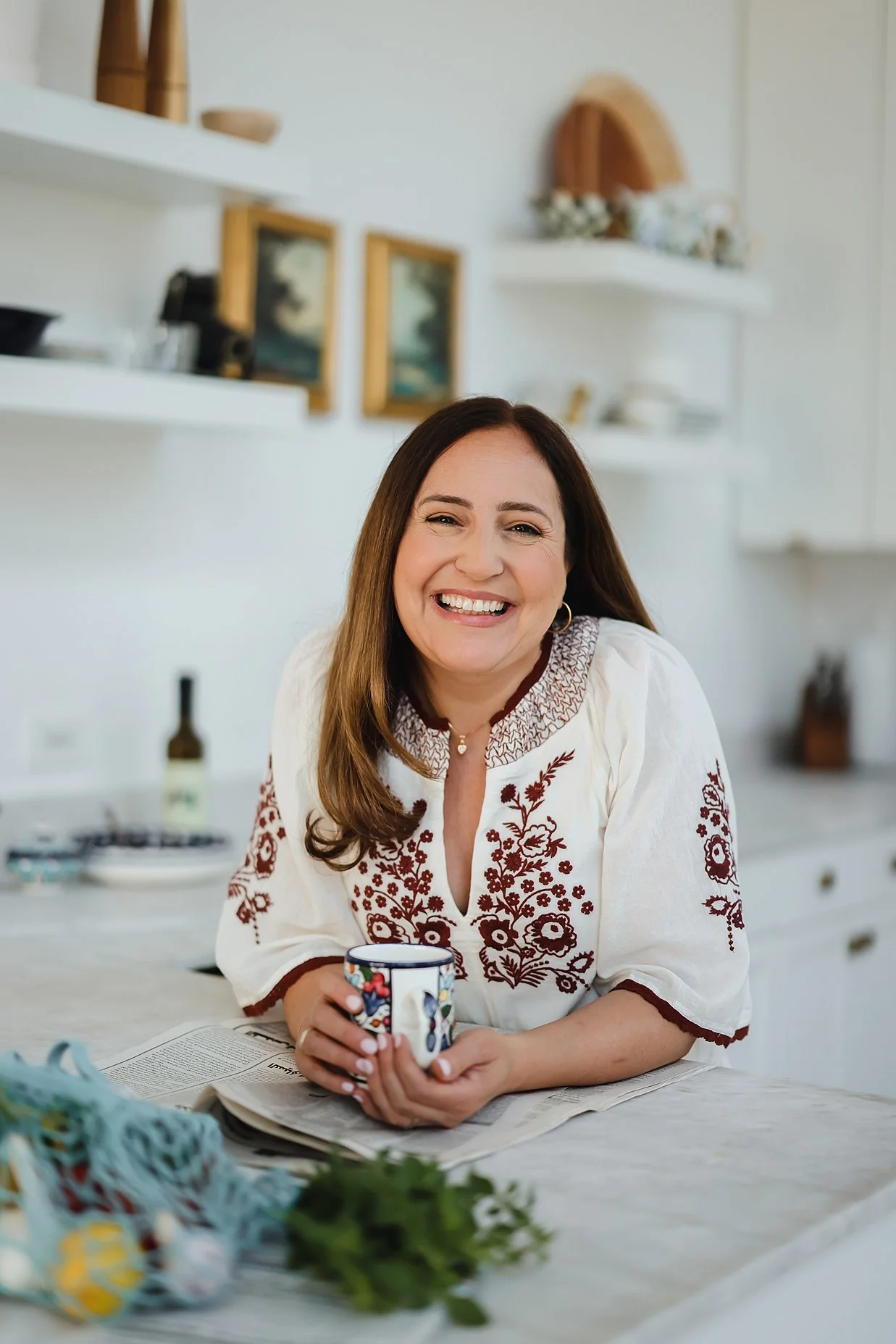 Smiling woman in a kitchen with a floral embroidered blouse, holding a colorful mug.