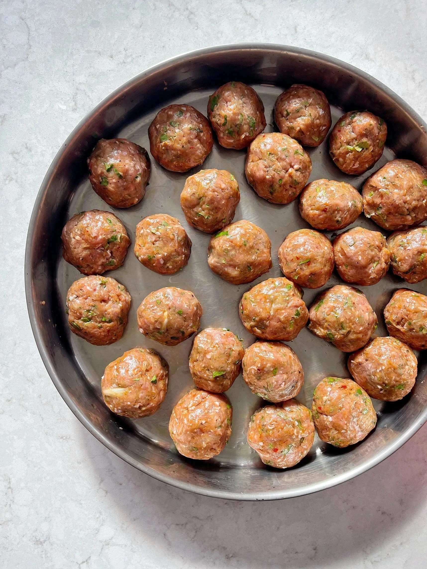 Raw meatballs with herbs in a round baking tray on a light countertop.