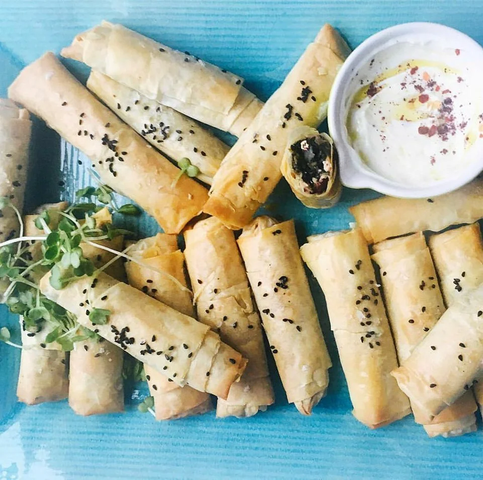 Plate of crispy rolled pastries with black seeds, served with a side of creamy yogurt dip on a blue surface. Fresh greens garnish.