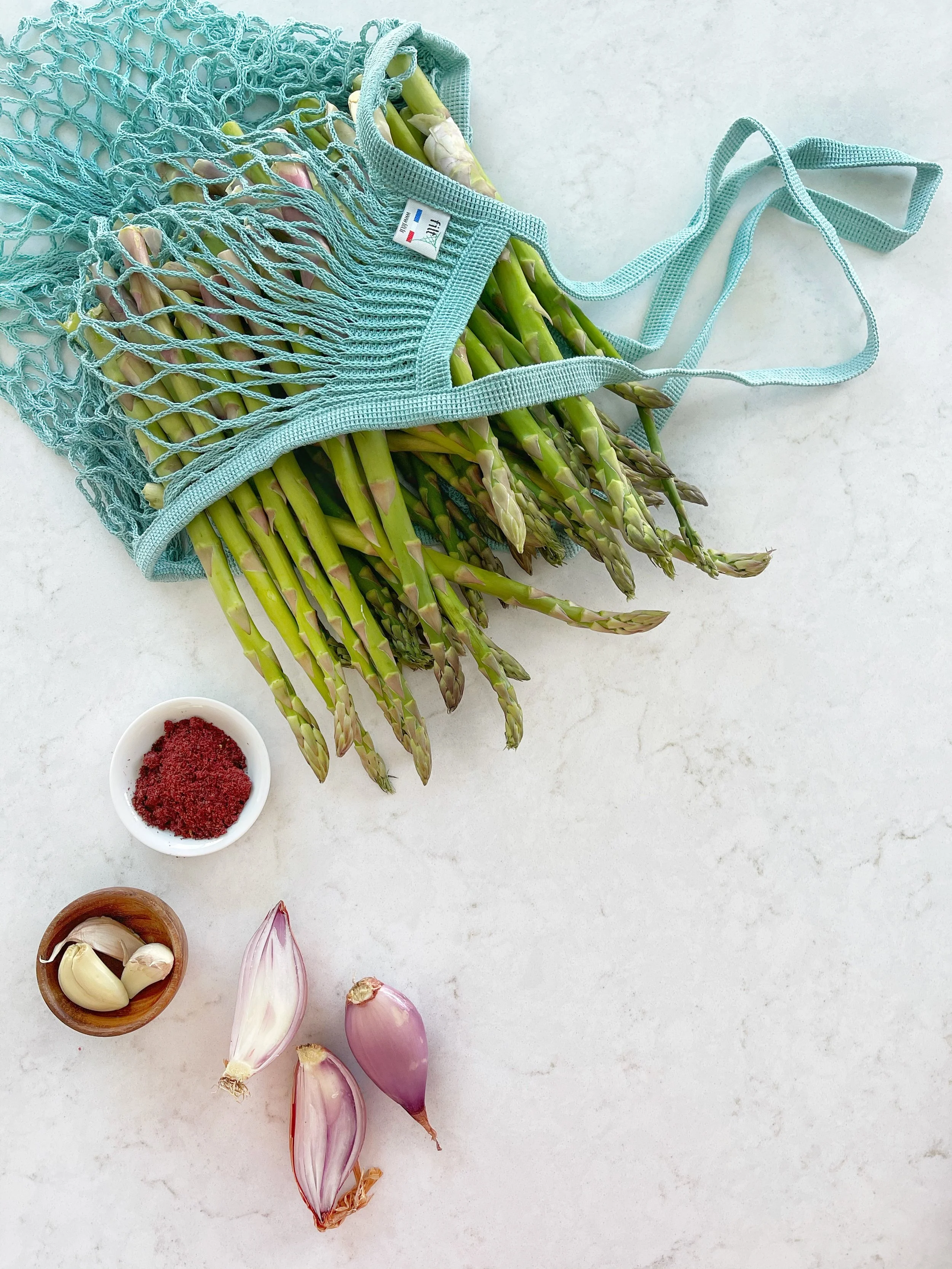 Fresh asparagus in a blue mesh bag, garlic cloves in a small wooden bowl, shallots, and a white bowl with spices on a white marble surface.