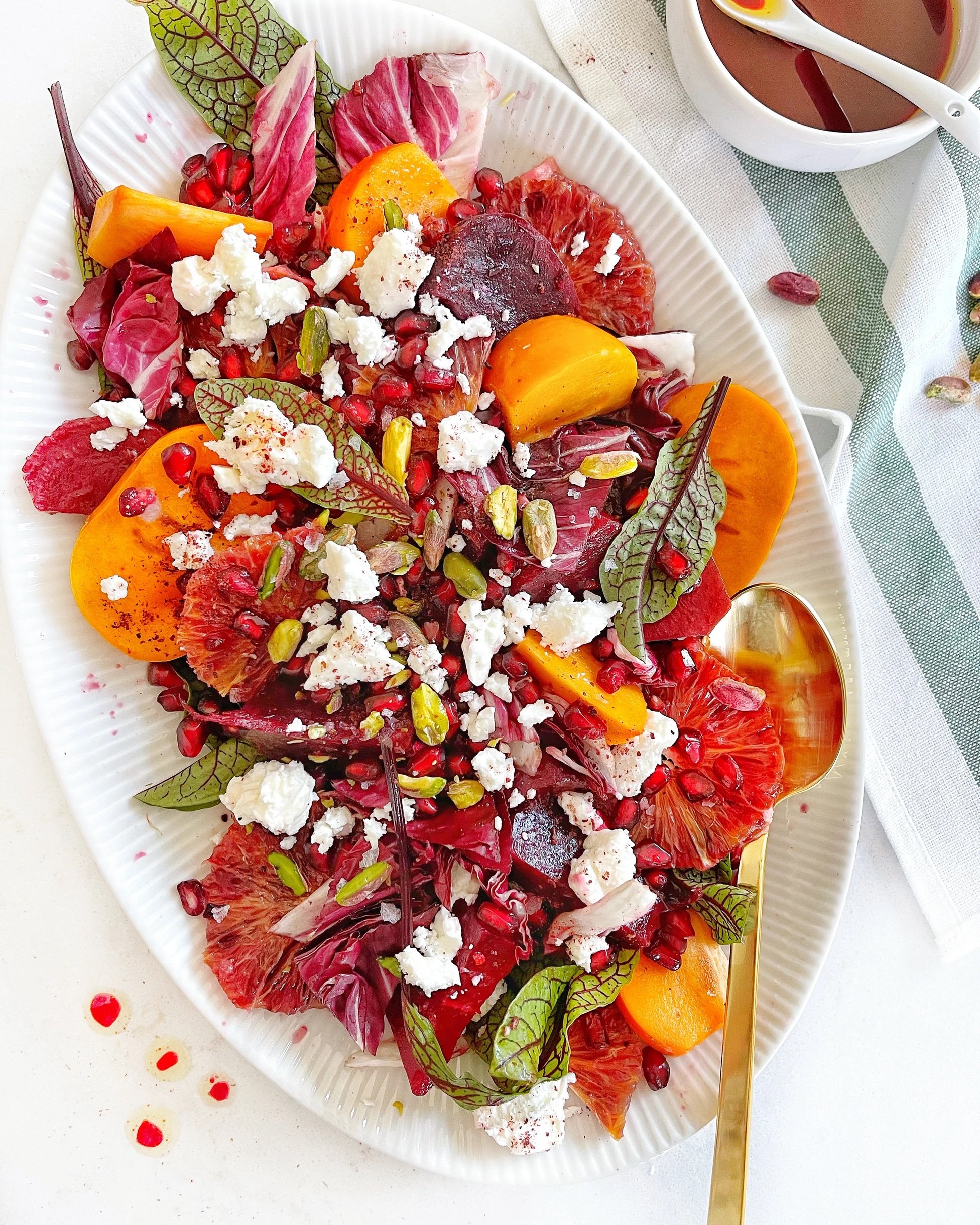 Colorful salad with orange slices, beetroot, mixed greens, pomegranate seeds, pistachios, and goat cheese on a white plate with a gold spoon.