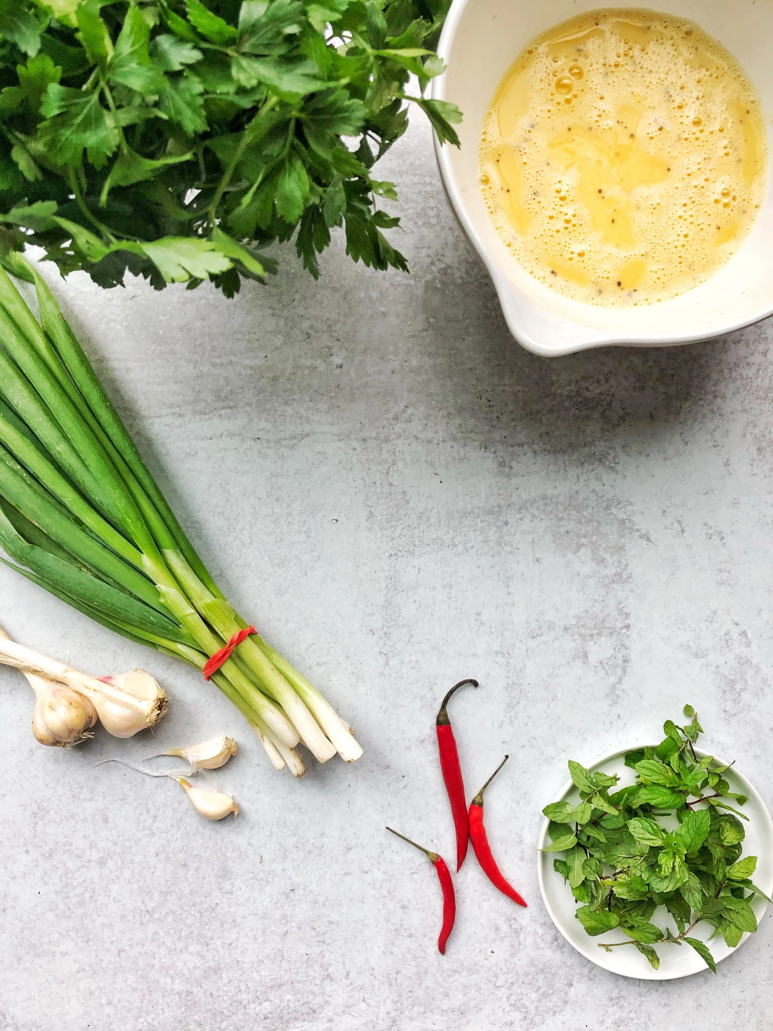 Fresh ingredients including parsley, green onions, garlic, red chili peppers, mint leaves, and a bowl of beaten eggs on a gray background.