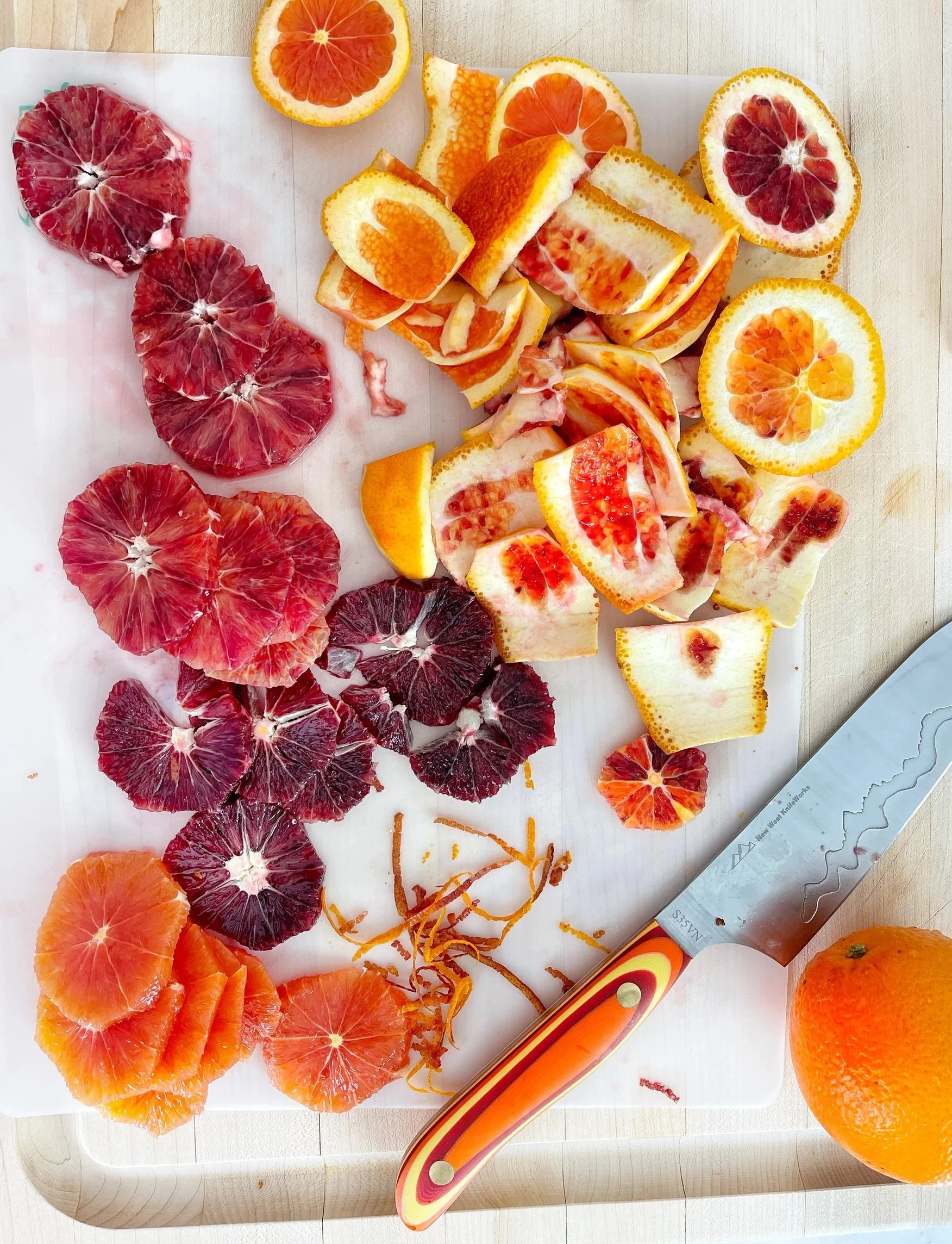 Slices and peels of blood oranges on a cutting board with a knife.