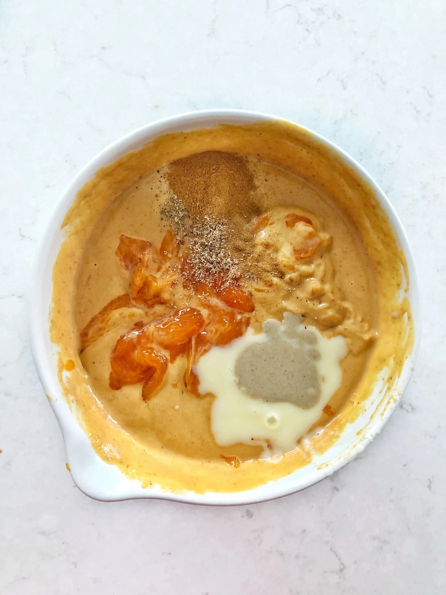 Bowl of pumpkin cheesecake batter with spices and cream on a white surface.
