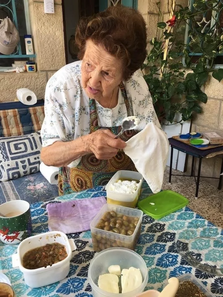 Elderly woman in apron holding a spoon, seated at a table with food containers including olives, cheese, and condiments on a patterned tablecloth. Outdoor setting with plants and a stone wall in the background.