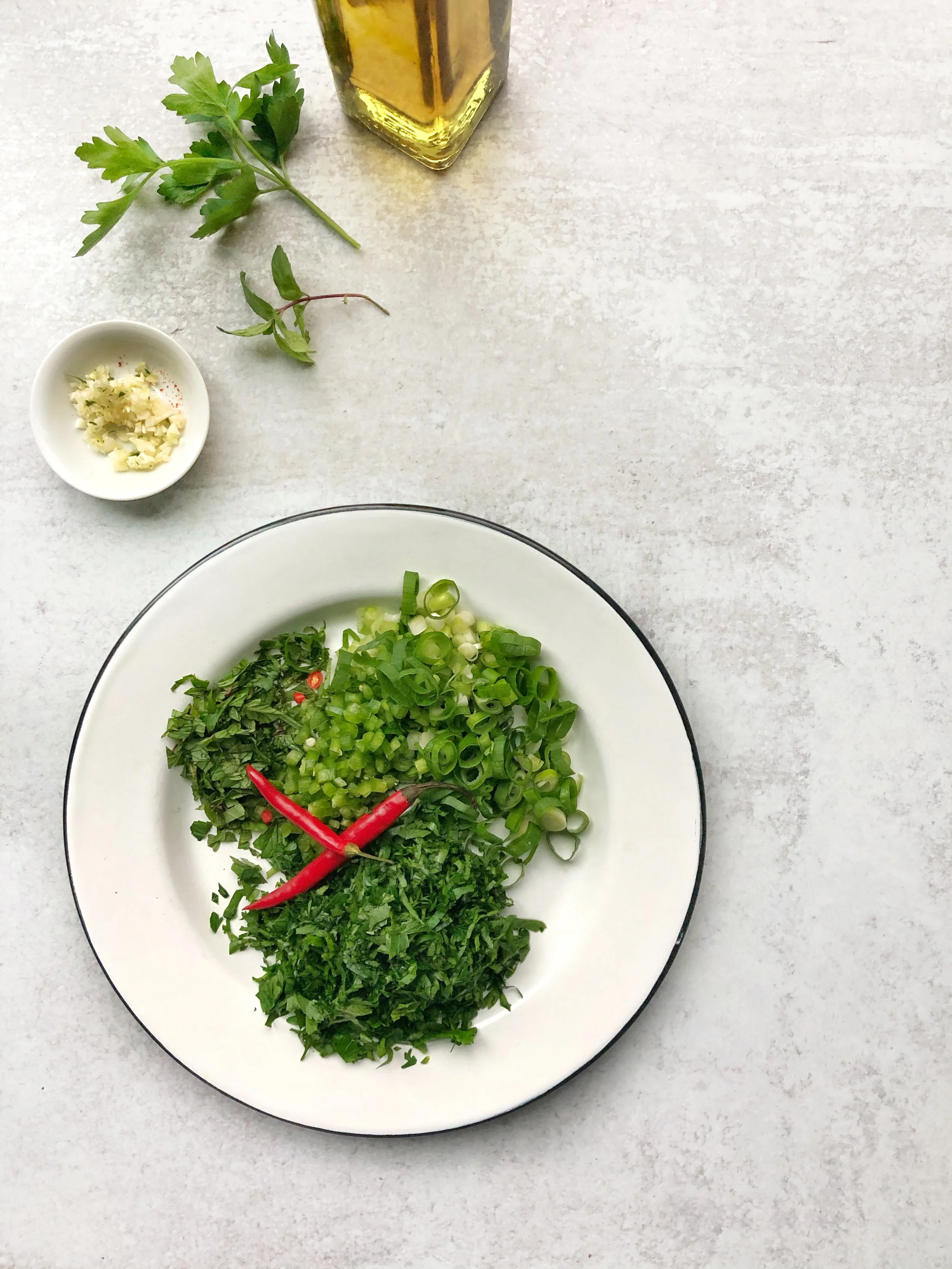 Herbs and spices on a white plate with green onions, chopped parsley, green chili, and red chili peppers, next to a small bowl of minced garlic and a bottle of olive oil on a light surface.
