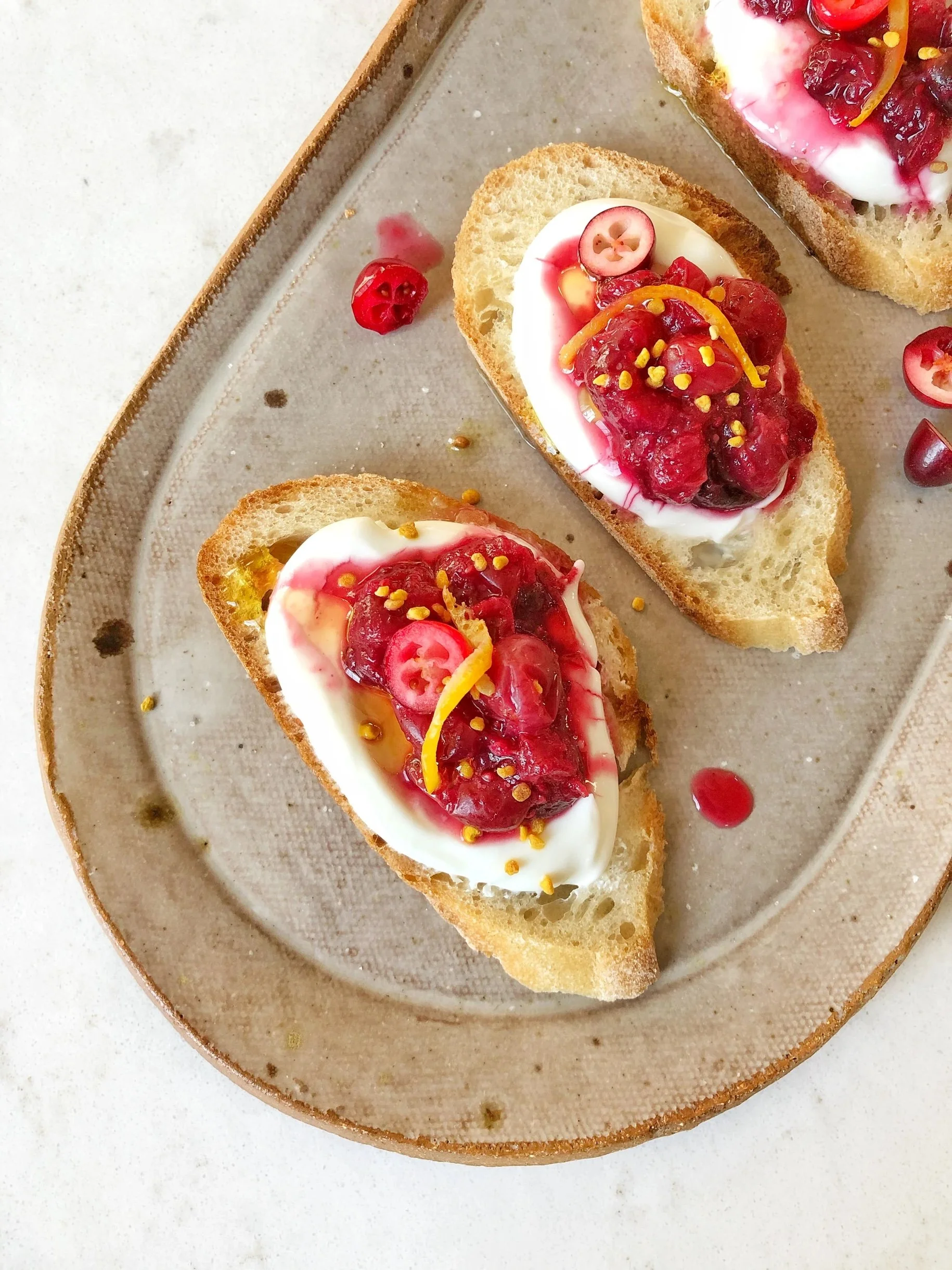 Slices of bread topped with cream cheese, cranberries, and orange zest, served on a beige plate.