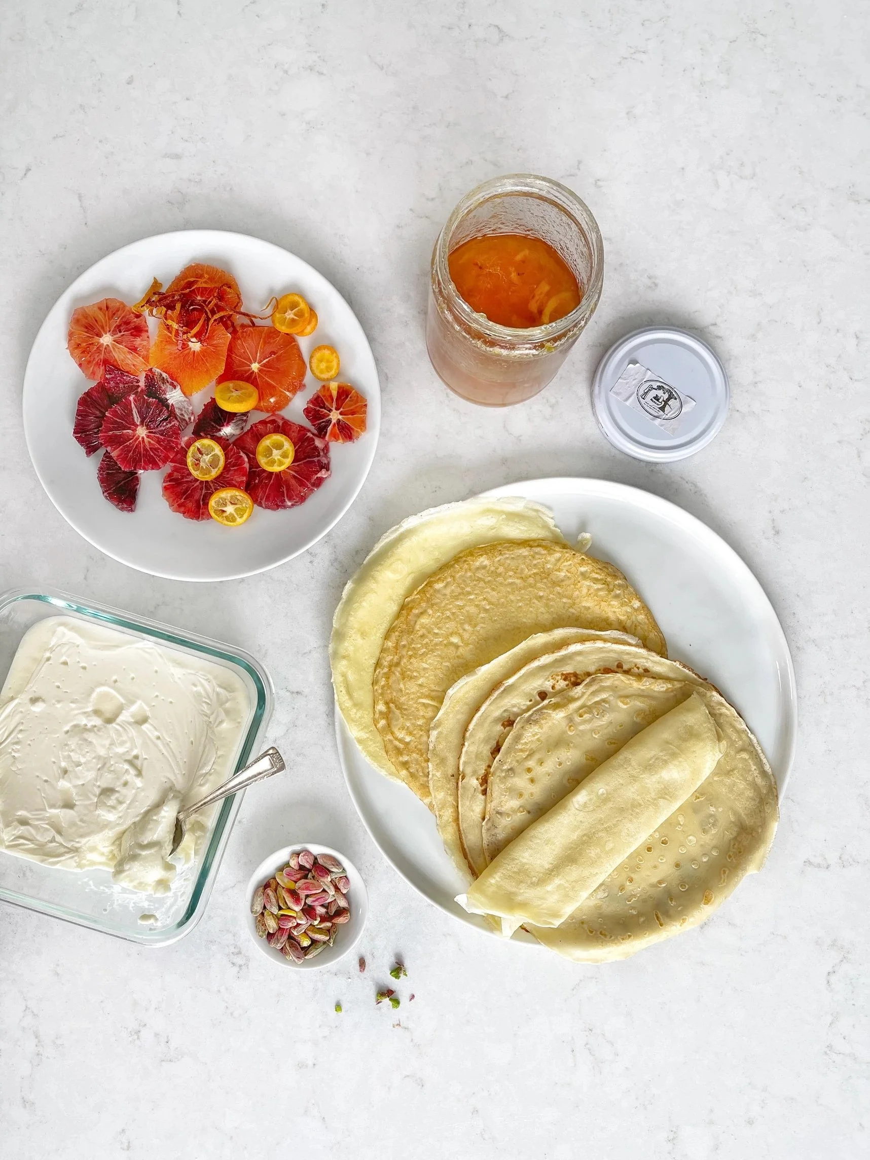 A plate of crepes with a jar of orange marmalade, a dish of cream, a small bowl of pistachios, and a plate of sliced blood oranges and kumquats on a light countertop.