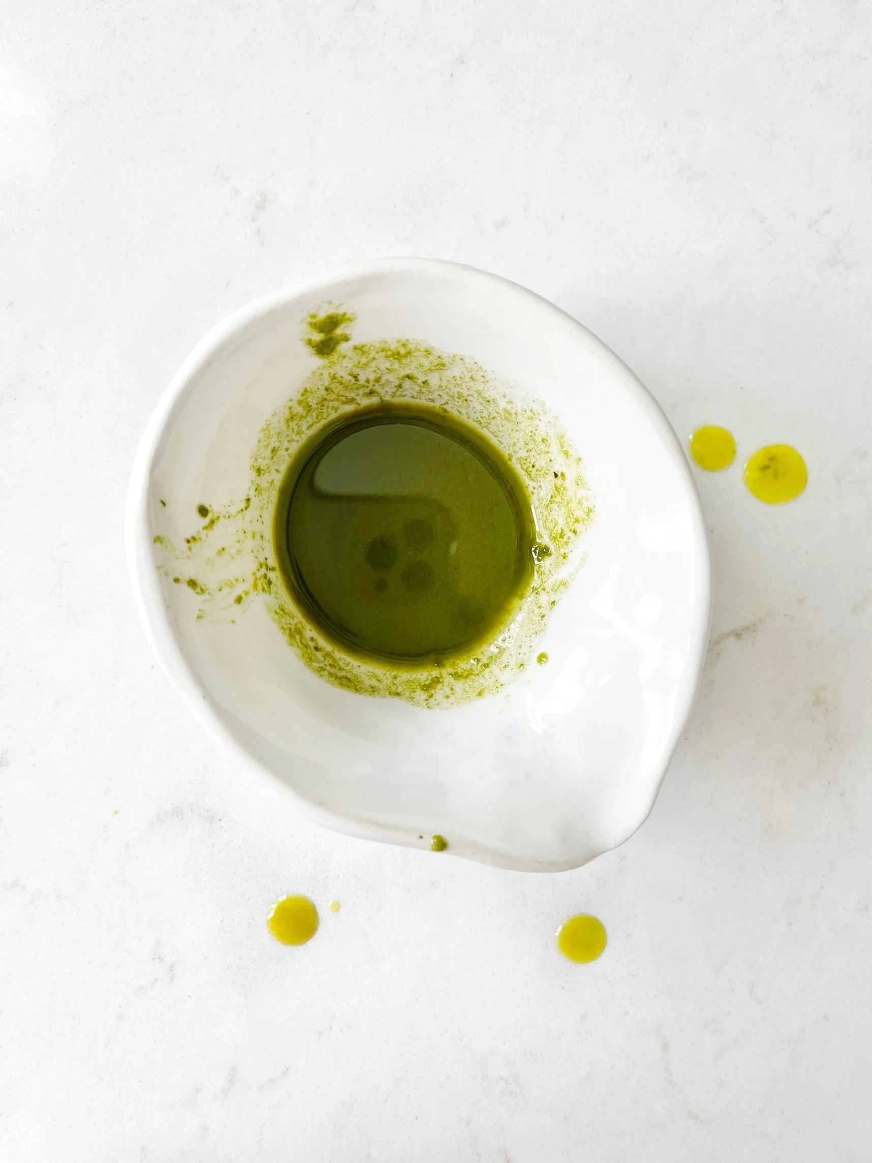 White bowl with green liquid on a marble surface, small green droplets nearby.