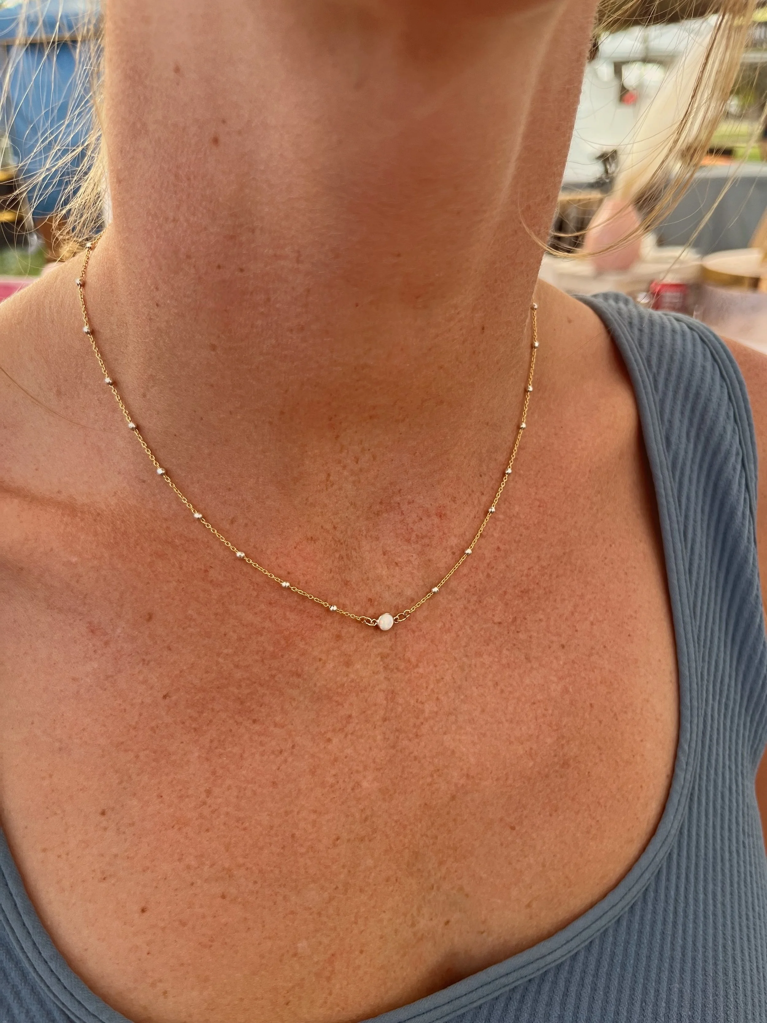 Close-up of a woman's neck and collarbone wearing a delicate gold necklace with tiny beads and a single pearl in the center. She is wearing a blue sleeveless top and has light skin with freckles.