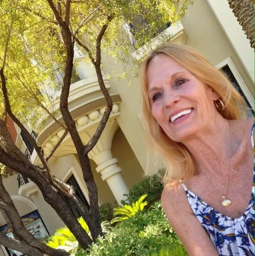 A smiling middle-aged woman with blonde hair wearing a sleeveless floral top and a heart-shaped pendant necklace, outdoors in front of a building with a rounded balcony and surrounded by greenery and trees.