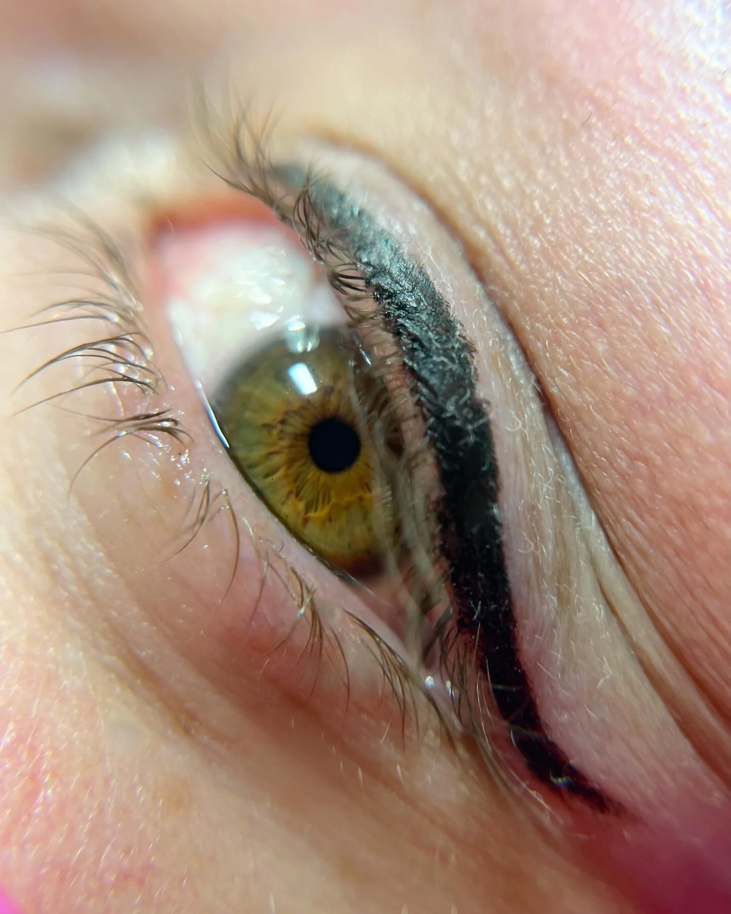 Close-up of a human eye with black eyeliner and light makeup, showing details of the yellow-green iris, eyelashes, and skin around the eye.
