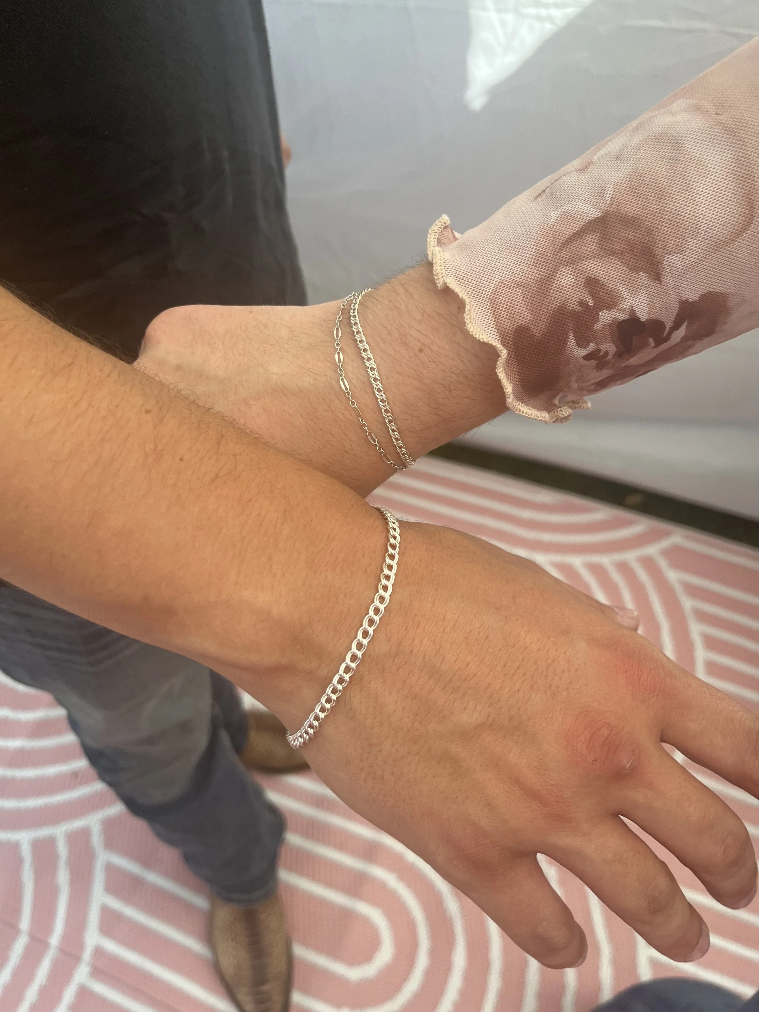 Close-up of a person's hands and wrists showing silver chain bracelets, with a pink floral sheer sleeve on one arm, standing on a pink and white patterned rug.