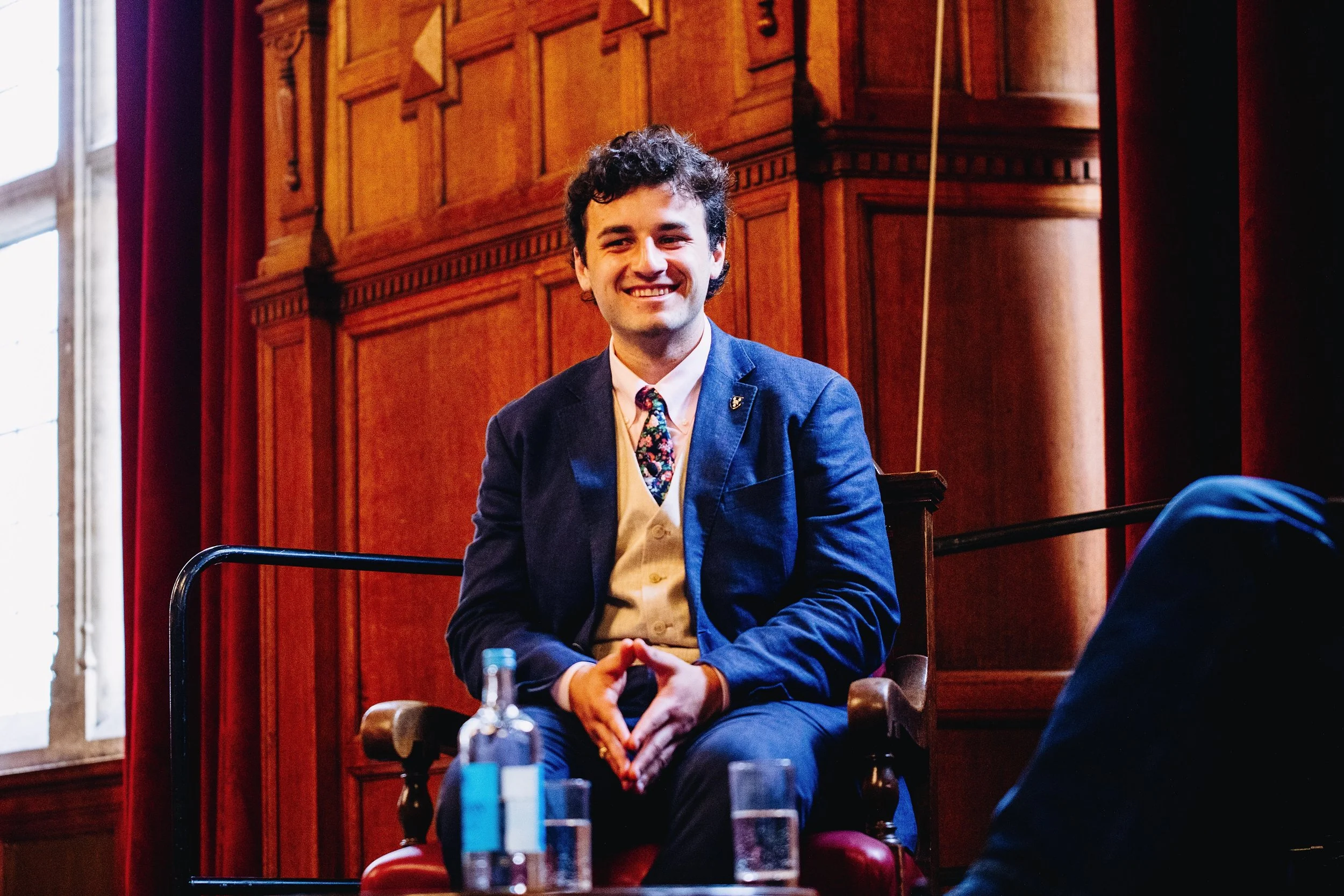 A young man with dark curly hair, wearing a blue blazer, cream vest, and floral tie, sitting in a wood-paneled room with red curtains, smiling, during a formal event or discussion.