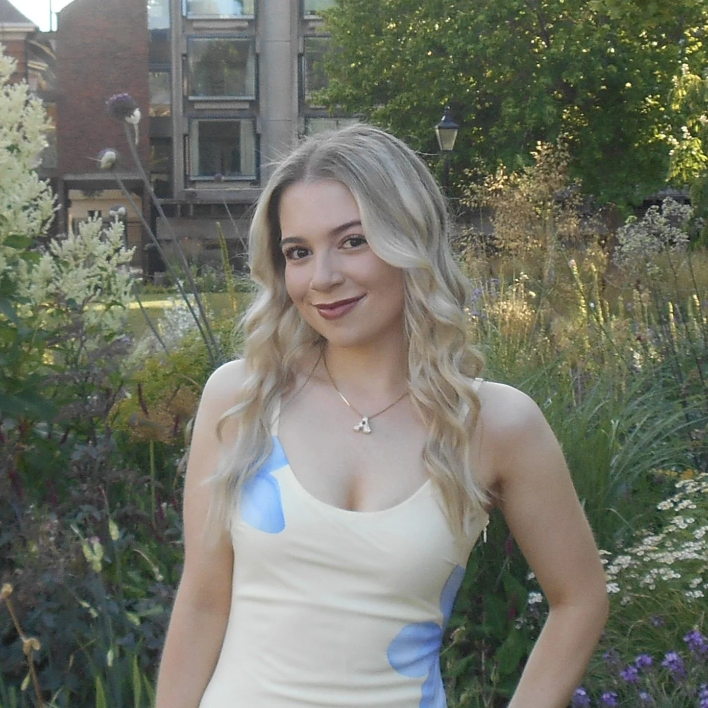 A young woman with long wavy blonde hair and fair skin, smiling and wearing a sleeveless dress with blue polka dots, standing in a garden with various flowering plants and greenery, and a building in the background.