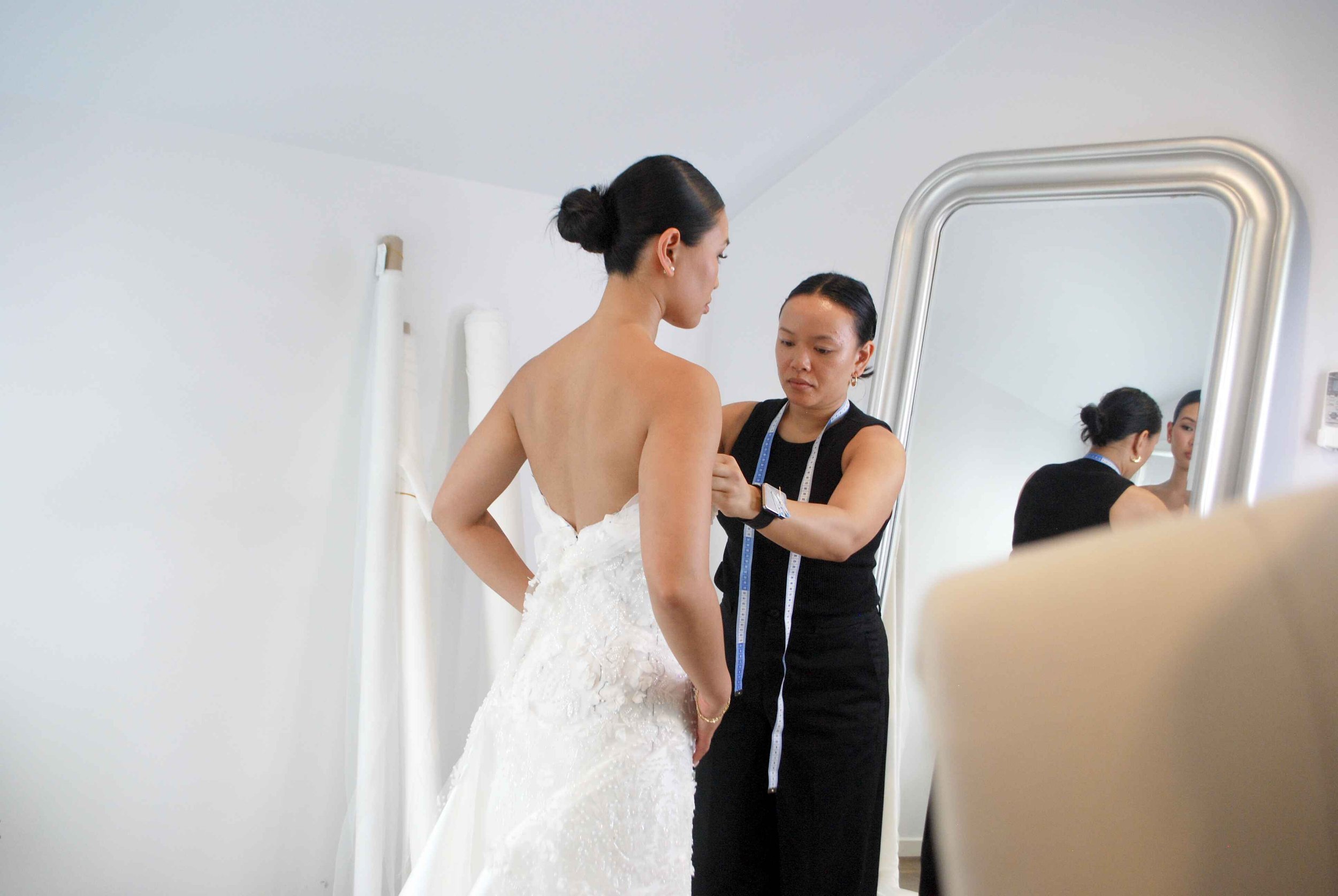 A fashion designer measures a bride in a strapless wedding gown in front of a mirror in a bright, minimalist studio.