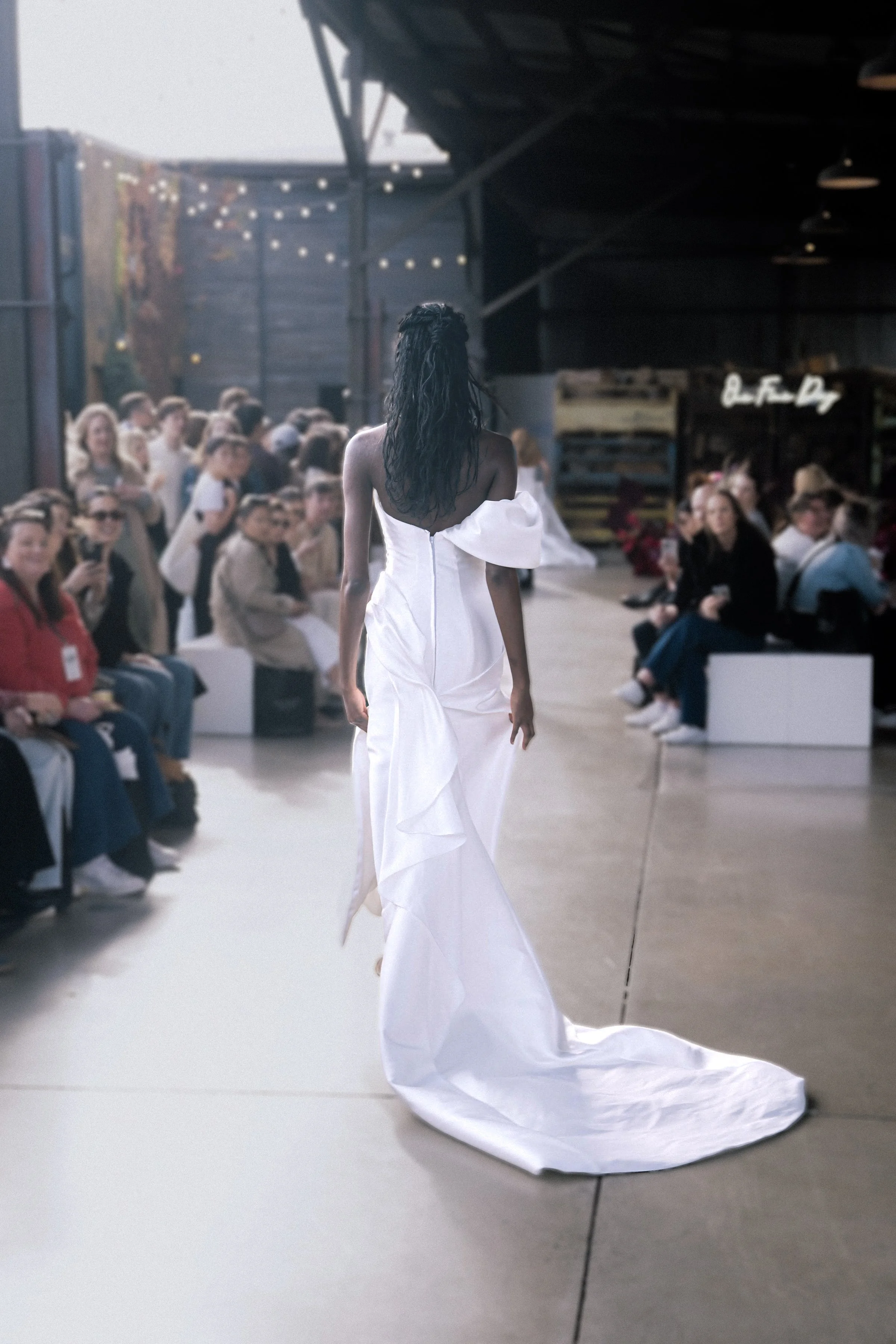 Model walks away on runway in a white gown with a long train and ruffled back detail, while audience watches attentively.