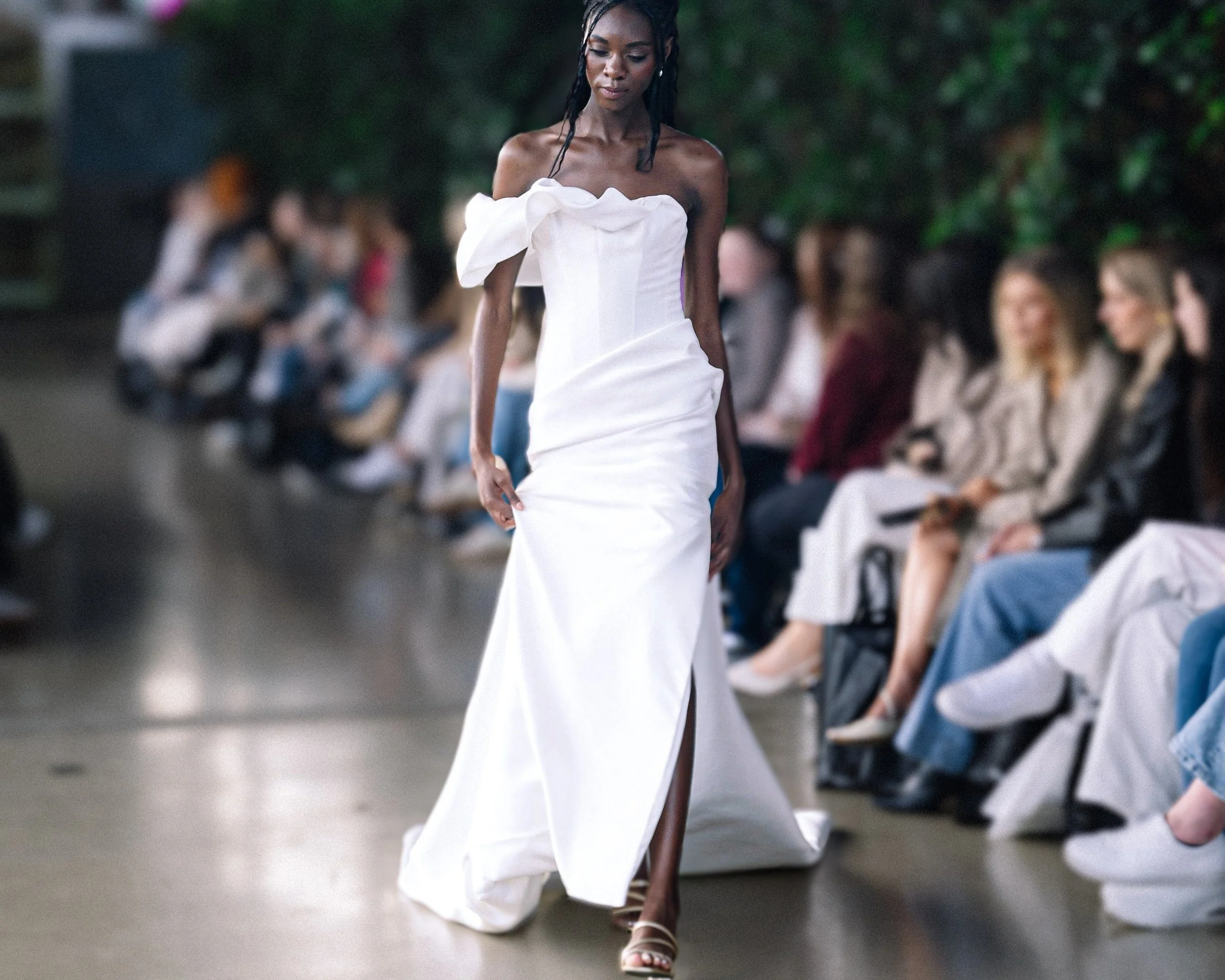 Model walks runway in an elegant off-shoulder white gown with a ruffled neckline and thigh-high slit, surrounded by seated audience.