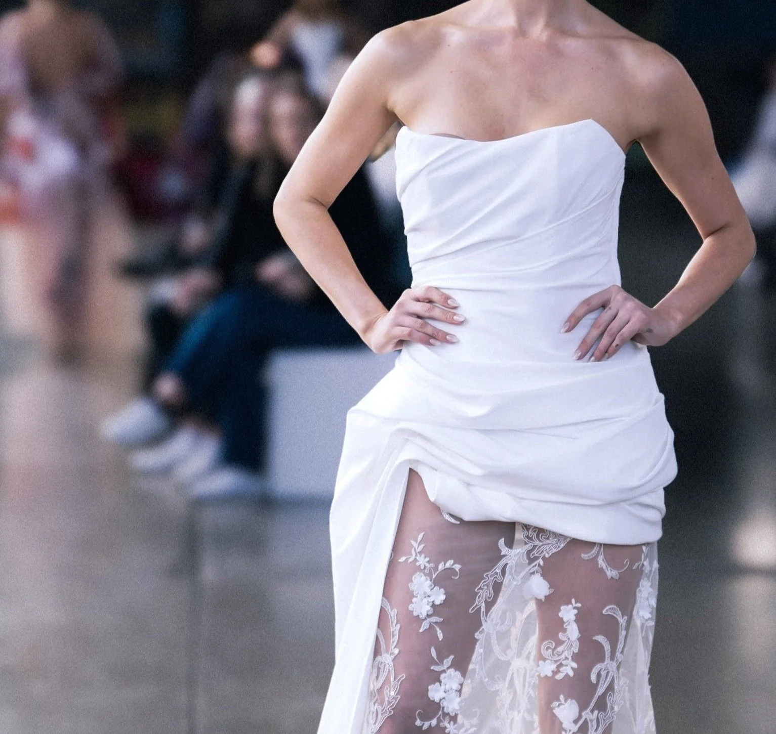 Close-up of a model wearing a strapless white dress with a ruched bodice and sheer floral-embroidered skirt on a runway.