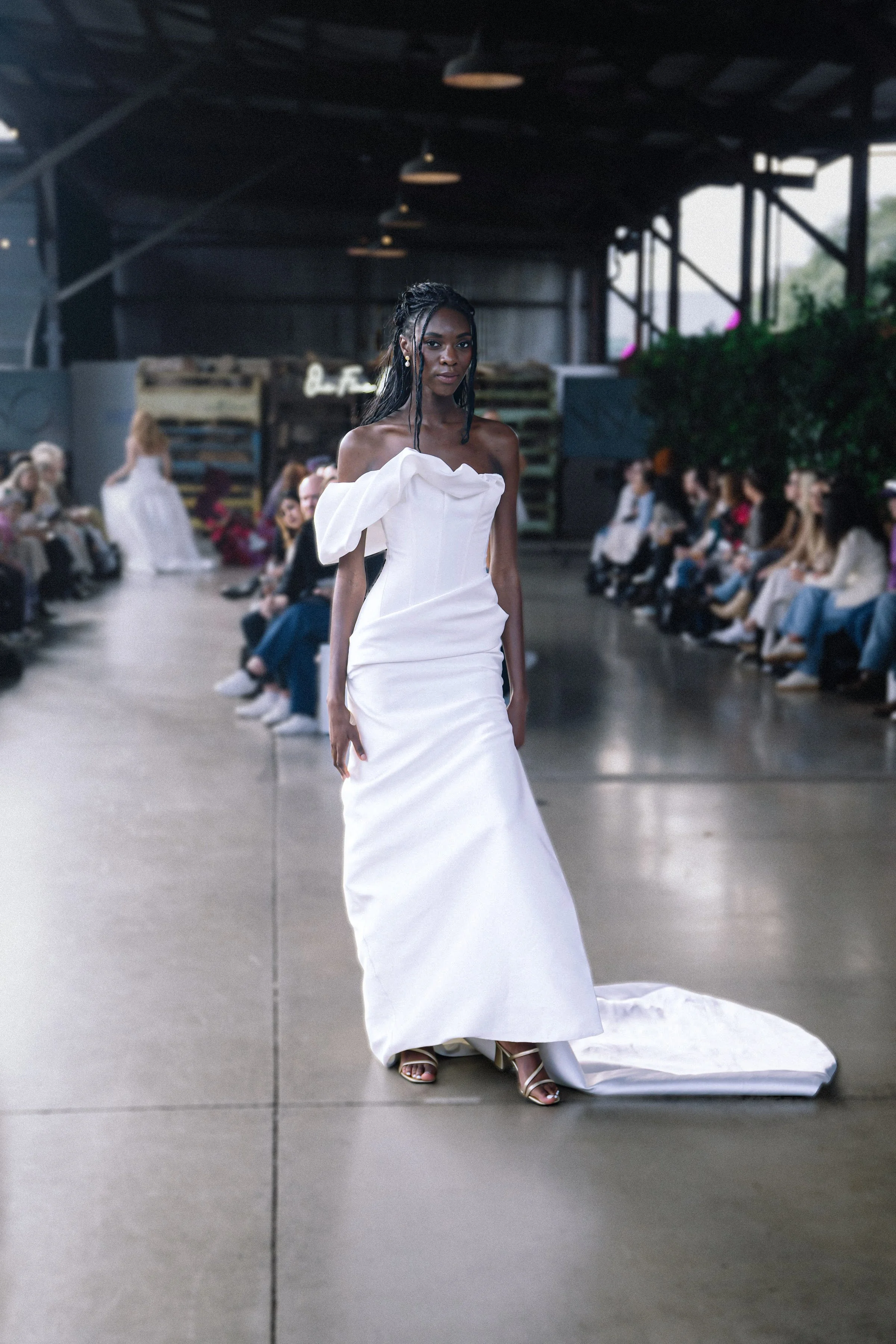 Model in a white off-shoulder gown with asymmetrical ruffle detail walks down the runway at an indoor fashion show.