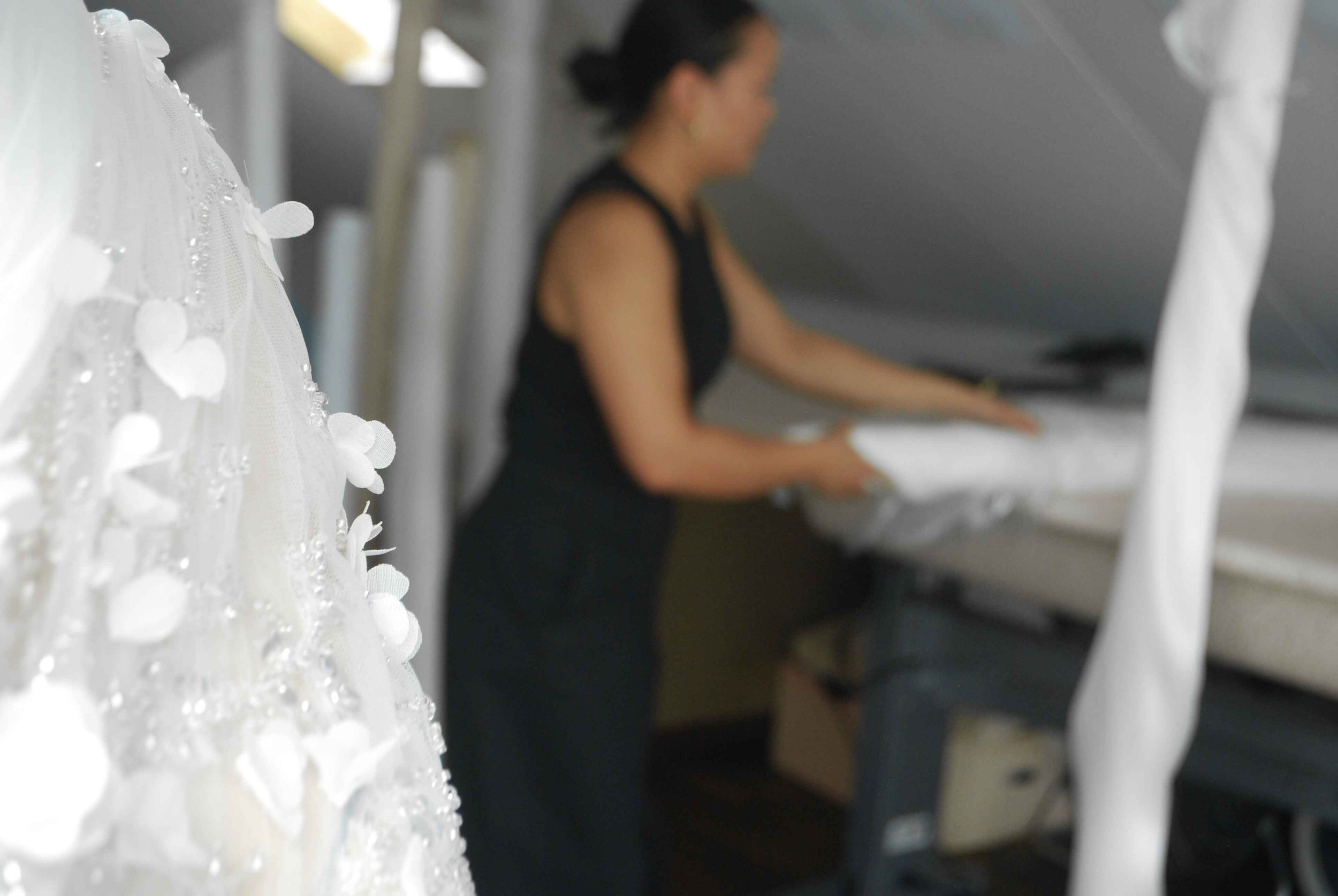 Close-up of a beaded wedding dress with floral details, with a blurred designer in the background folding white fabric.