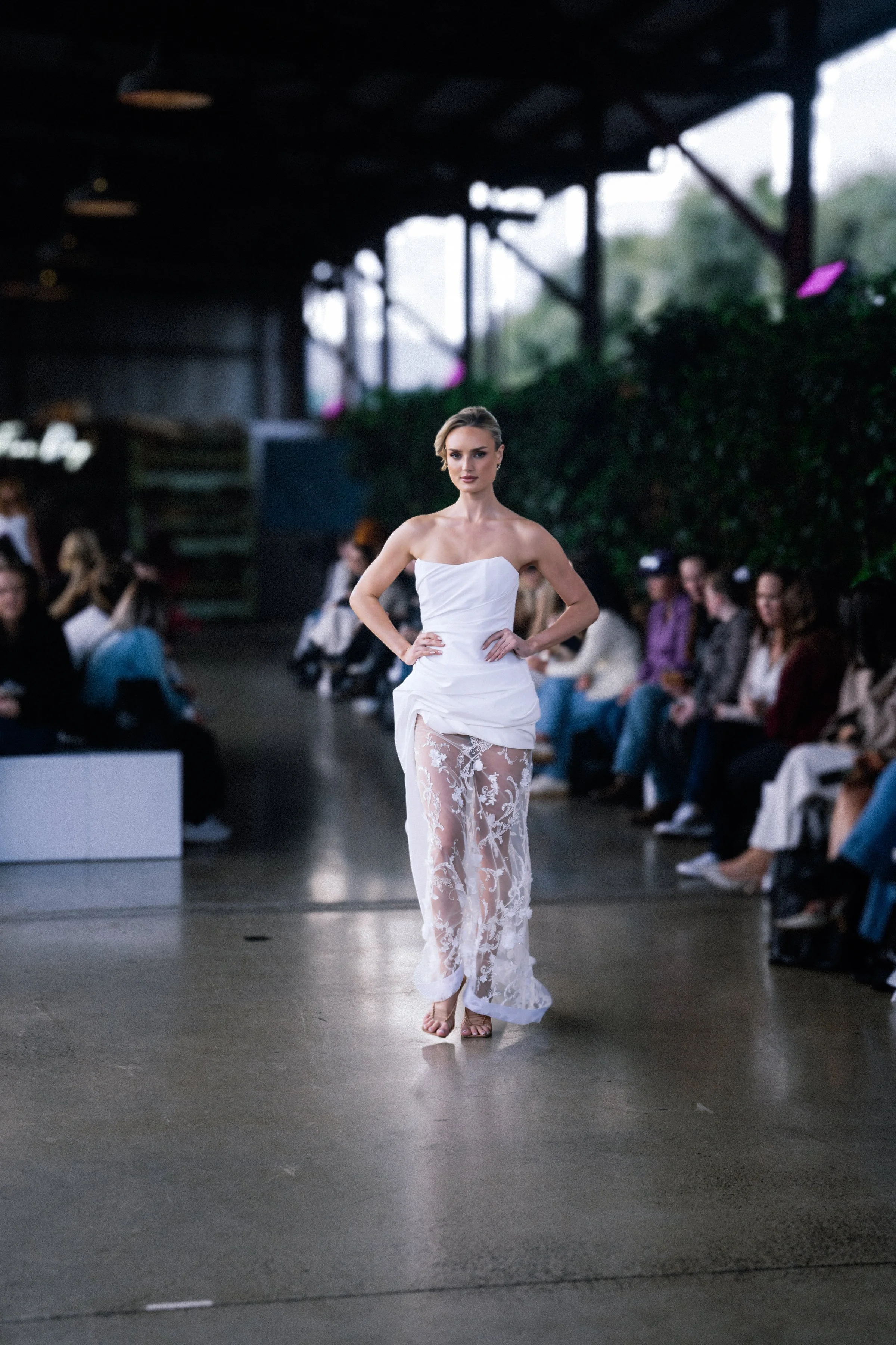 Model walks the runway in a strapless white gown with a ruched bodice and sheer floral-embroidered skirt at a fashion show.