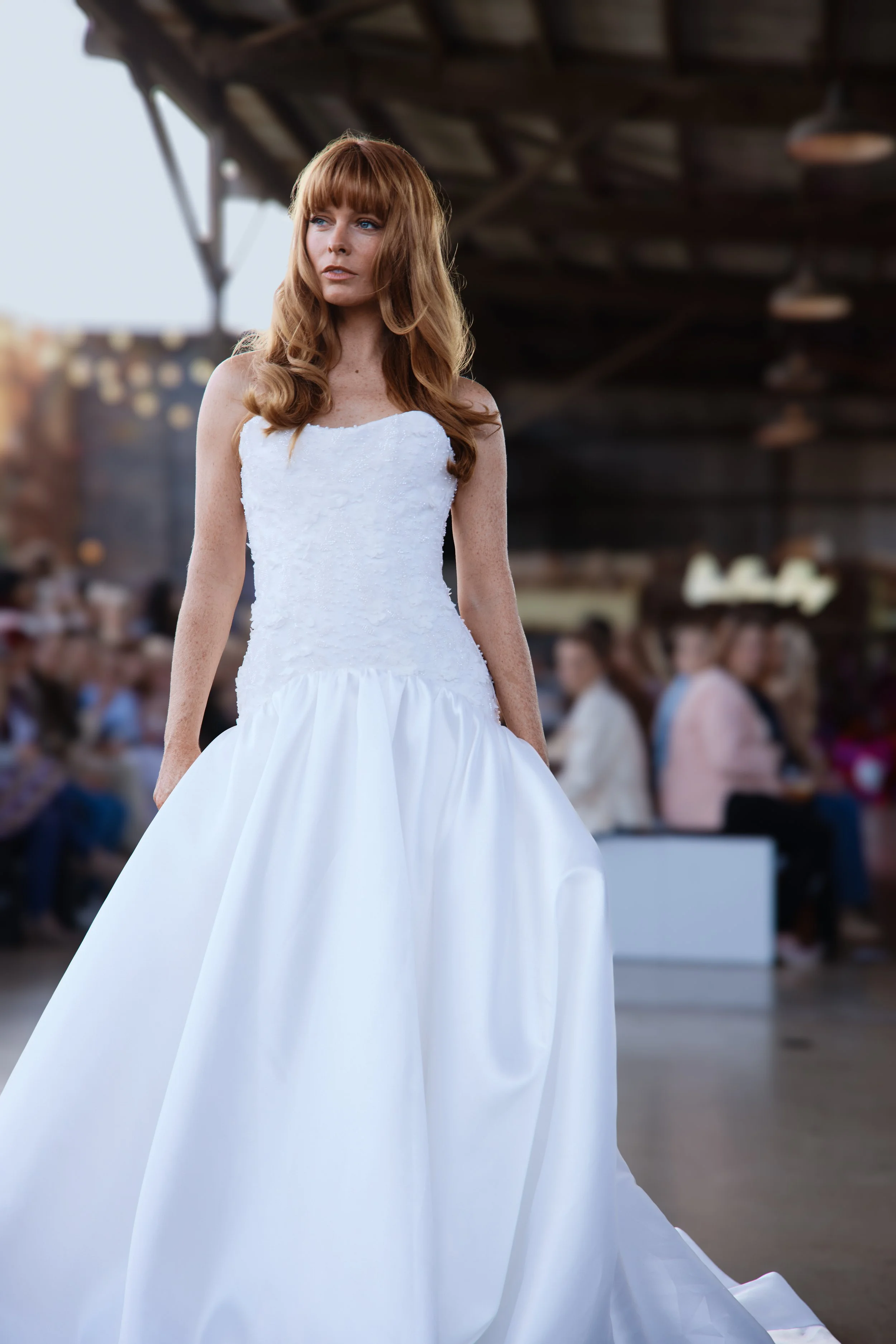 Model in a strapless white wedding gown with a full satin skirt walks a runway at a bridal fashion show, crowd seated behind her.